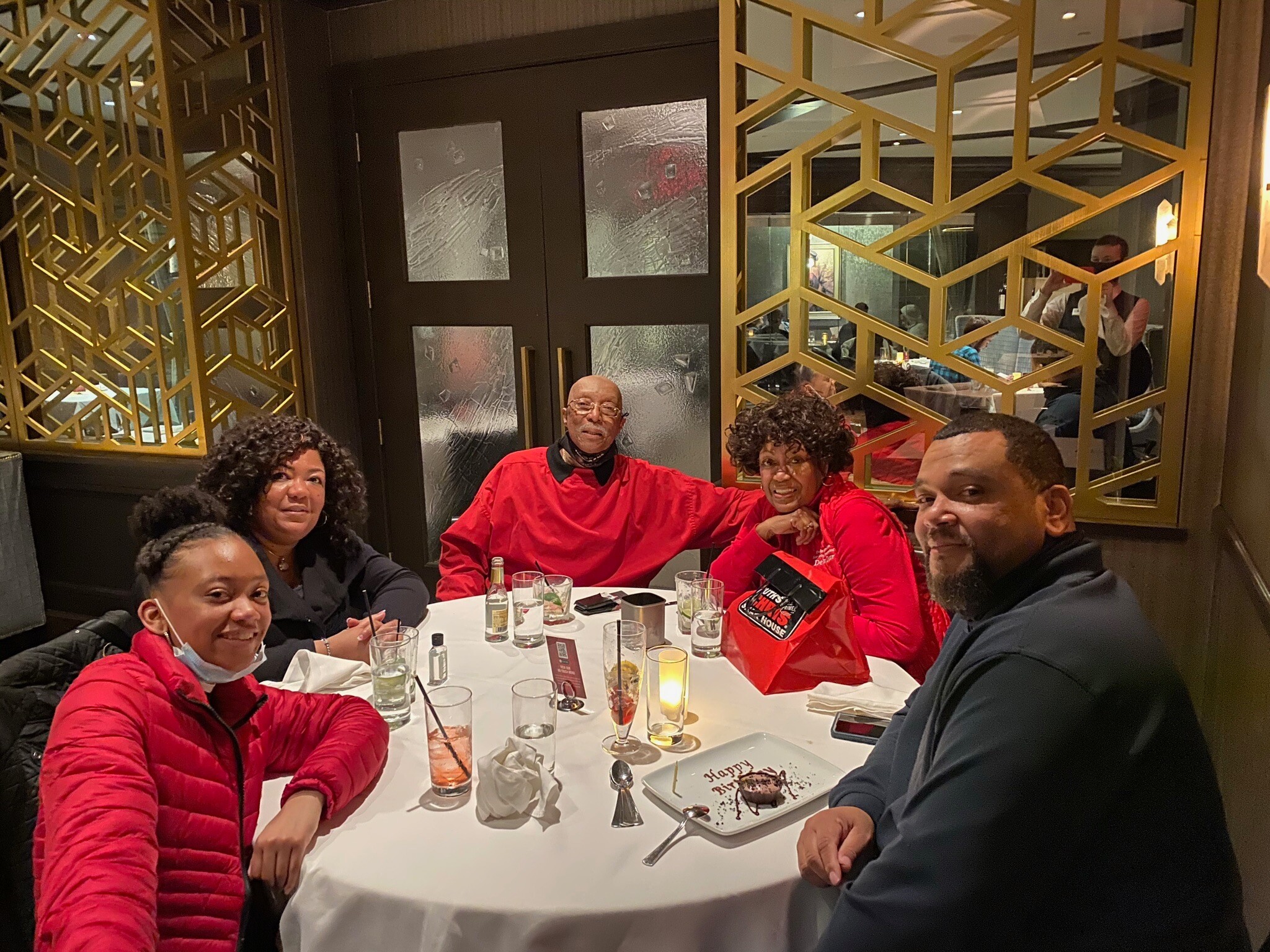 A group of friends sit around a restaurant table, taking a group selfie with birthday cake and drinks visible on the table.