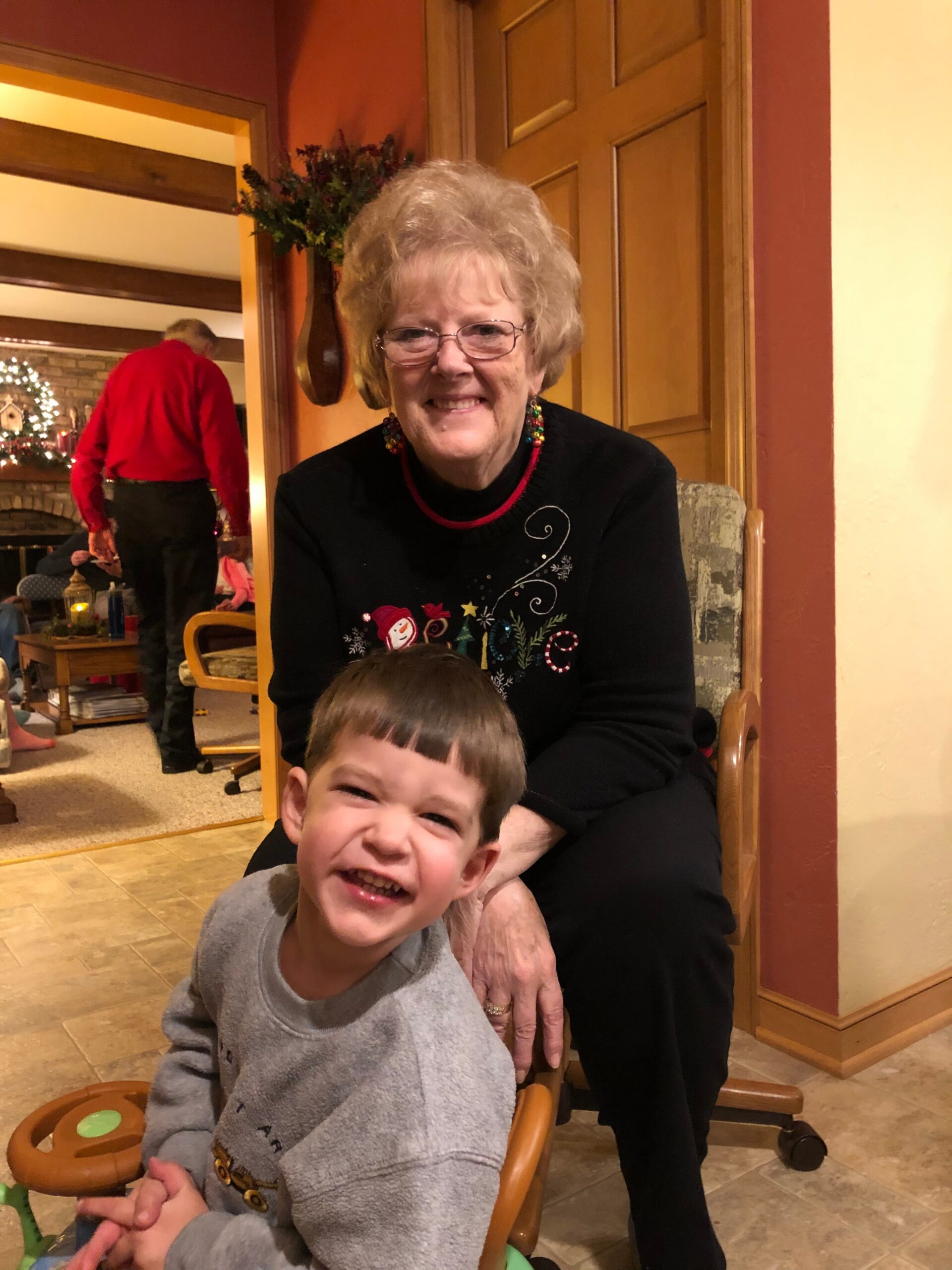 Smiling elderly woman with glasses and a festive sweater sits with a laughing young boy in a home Christmas gathering all indoors.