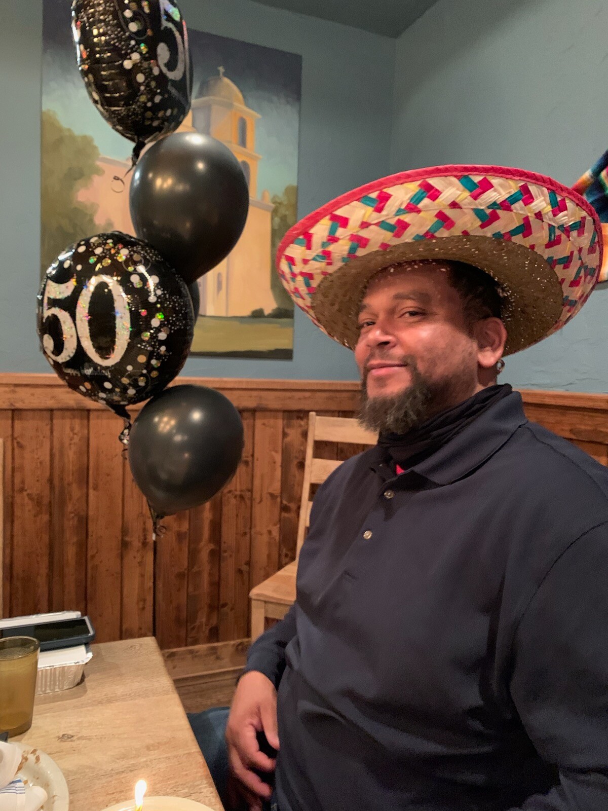 Man in a dark shirt wearing a colorful sombrero, celebrating a birthday at a wooden table with black balloons reading 50 in the background.