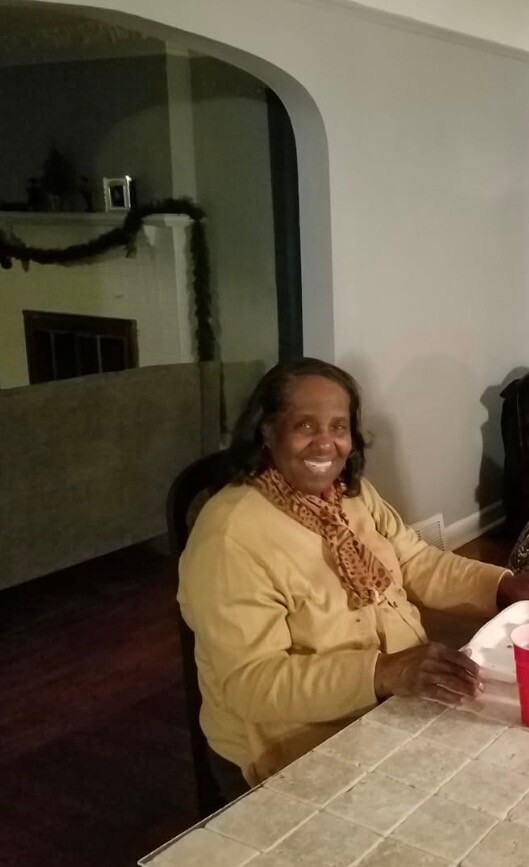 Smiling woman in a yellow sweater and patterned scarf sits at a tiled table indoors, with a garlanded mantle in the background.