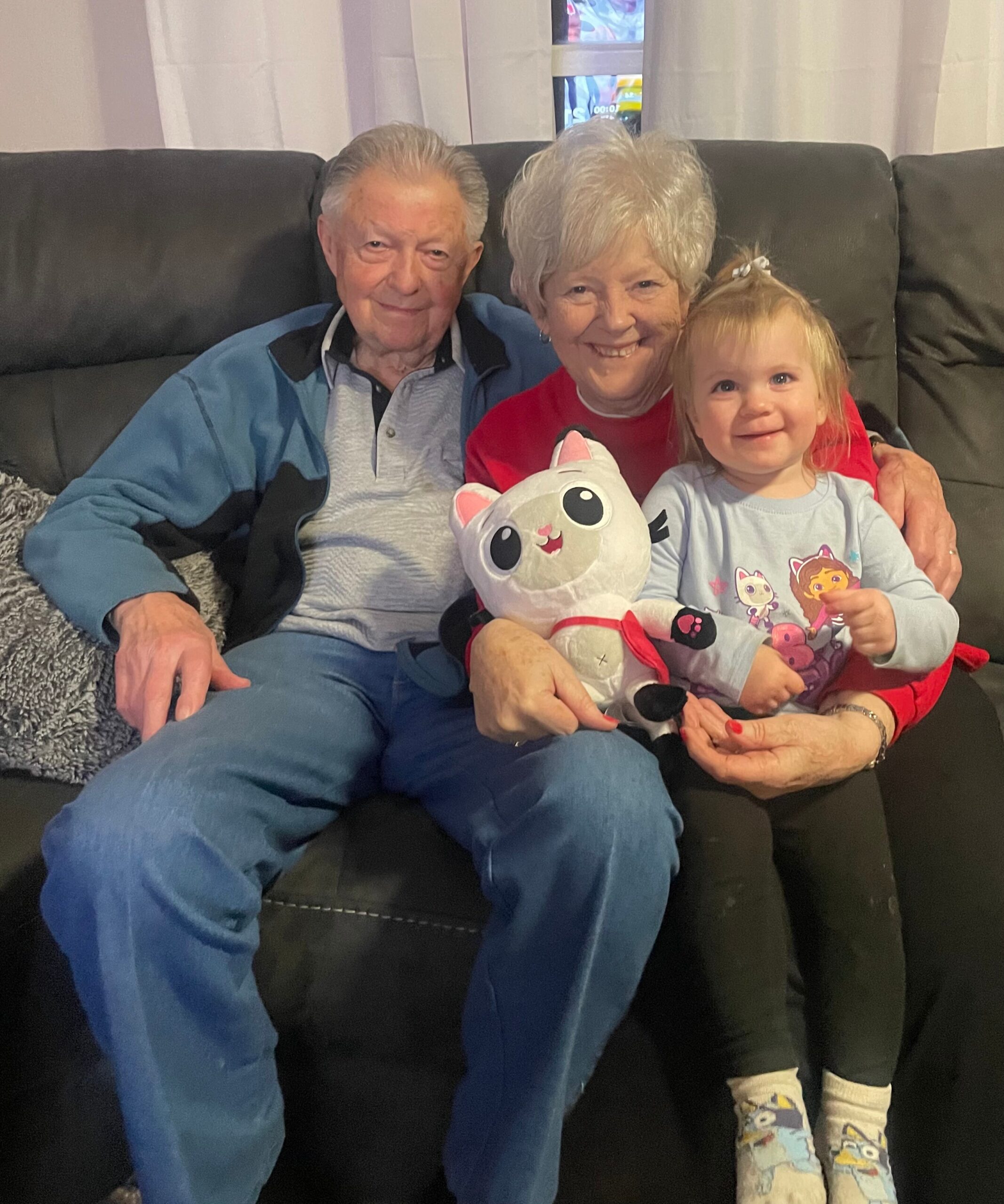 Grandfather, grandmother, and young girl sit together on a black sofa, smiling, with the girl holding a white plush cat toy with pink ears in her lap.