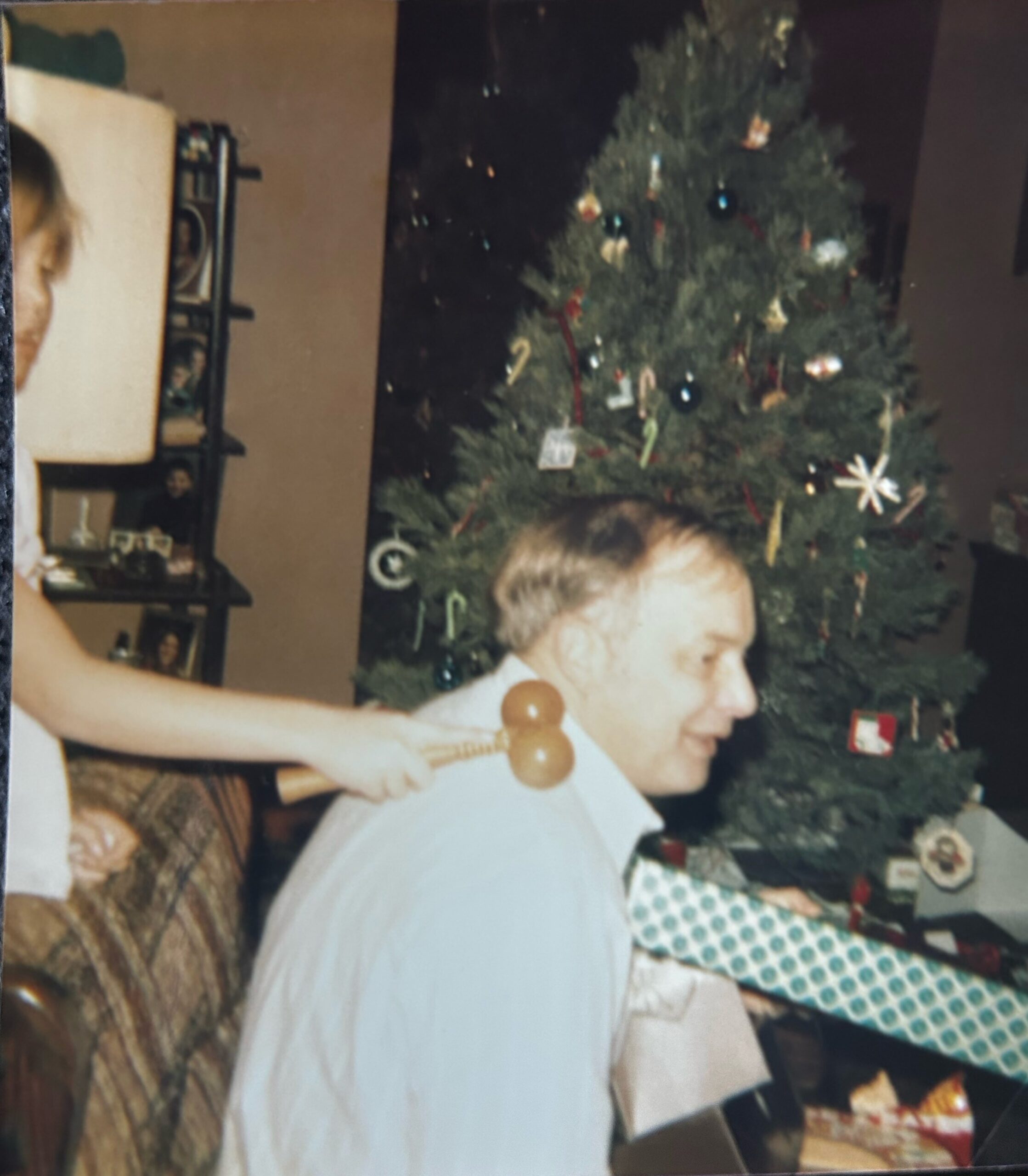 Man in a white shirt stands near a decorated Christmas tree with ornaments and wrapped gifts indoors, holding a wooden toy in front of him.