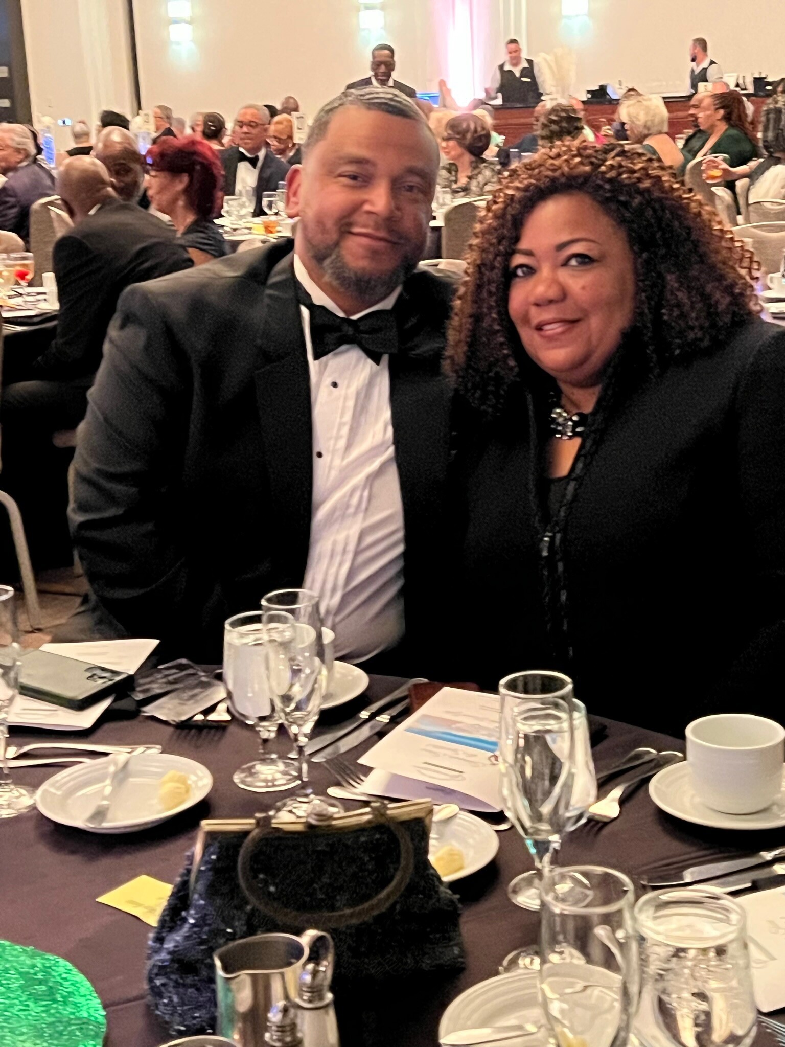 A smiling couple in formal wear seated at a banquet table during a gala event, with place settings and glasses around them.