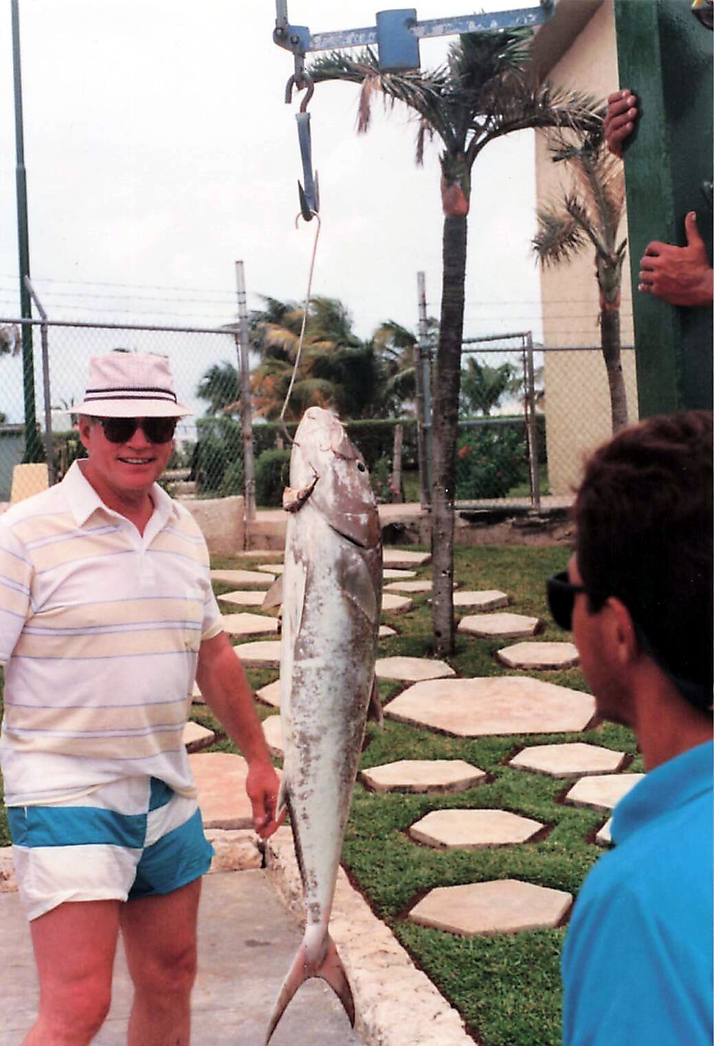 Man in a striped polo shirt, hat, and sunglasses stands beside a large fish hanging from a lifting mechanism outdoors.