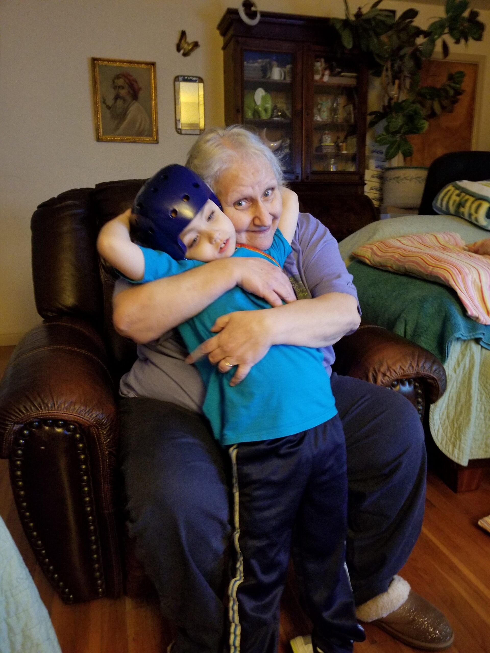Older woman sitting in a brown leather chair hugs a child in a blue shirt wearing a purple helmet indoors.