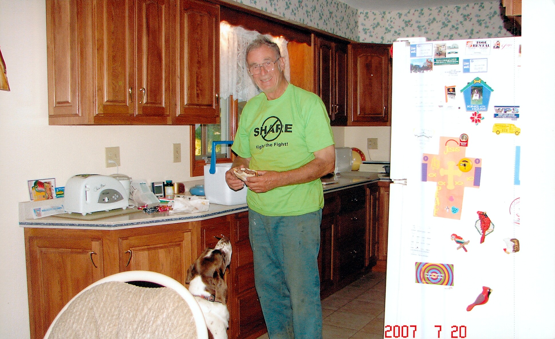 Man in a bright green shirt stands in a kitchen, smiling while holding a sandwich; a dog looks up at him.