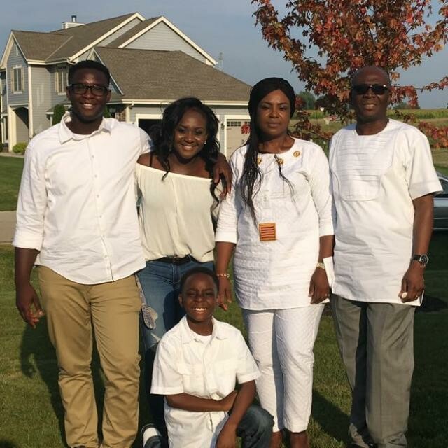 Family of five posing on a green lawn in front of a suburban house, all wearing white tops, smiling. Option 2: Five-member family outdoors, two adults and three children, standing together in white outfits in a sunny yard. Option 3: Group portrait of a family standing on a lawn with a beige house behind, all in white clothing.