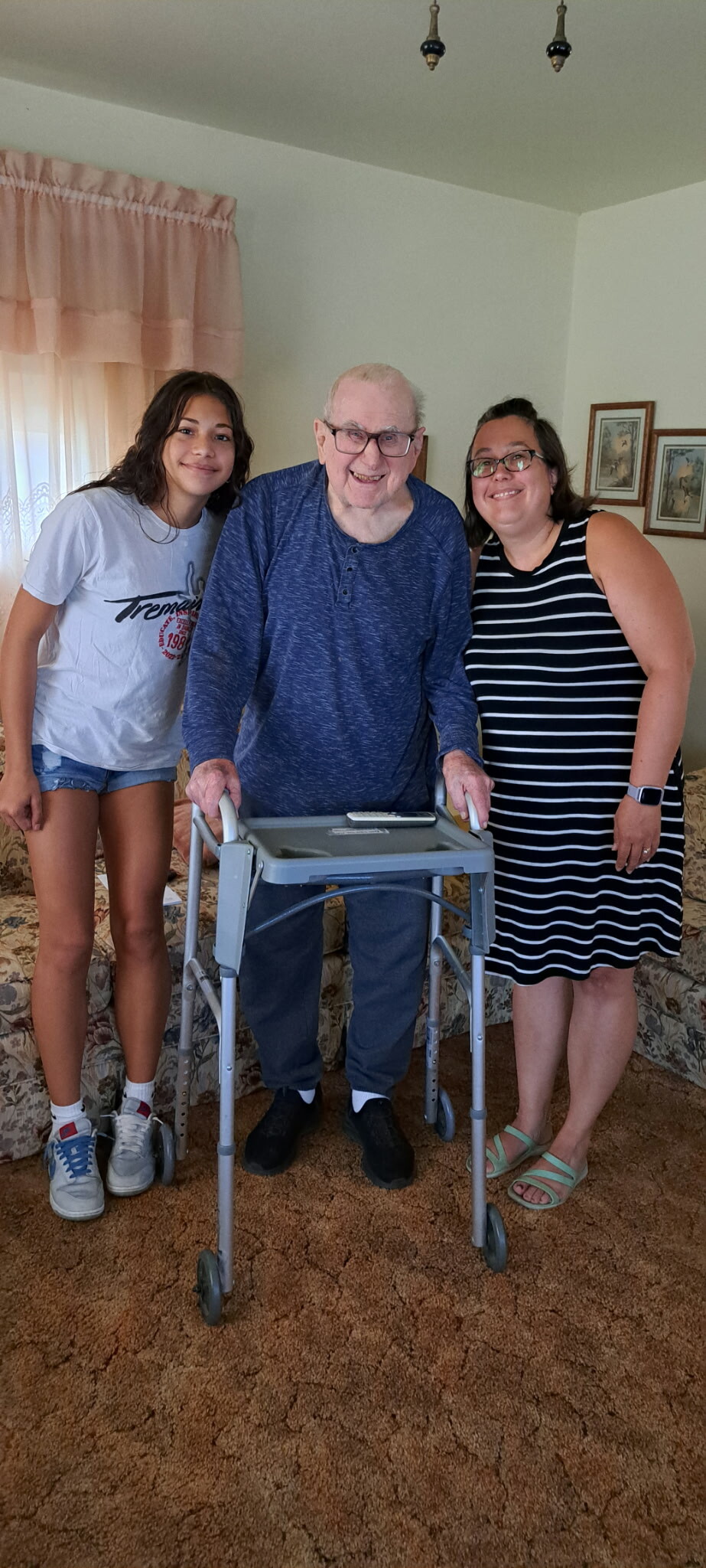 Three people posing for a photo at home: an elderly man using a walker, flanked by two smiling women.