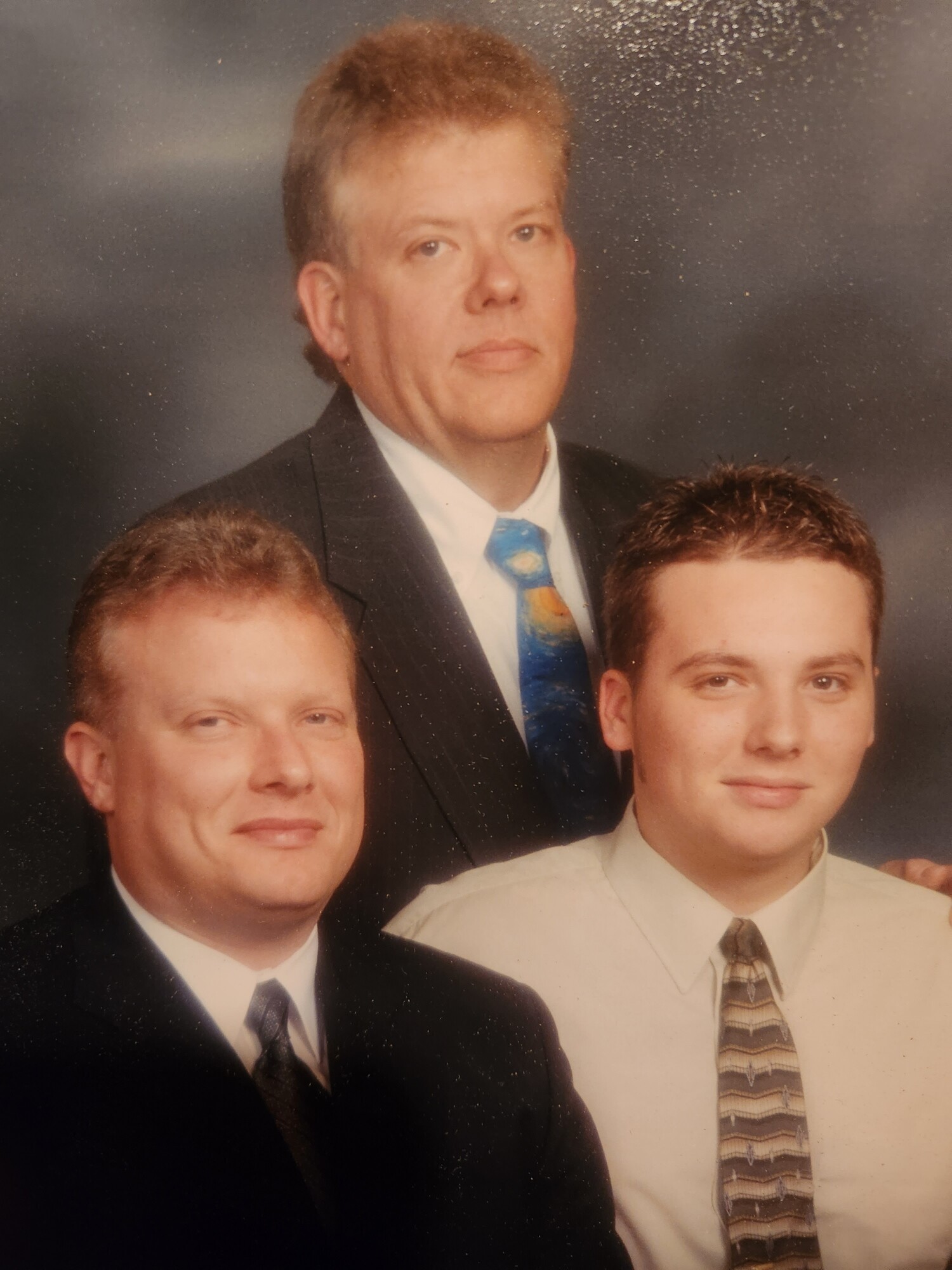 Group portrait of three men in suits posing in a studio backdrop; centered man wears a blue patterned tie.