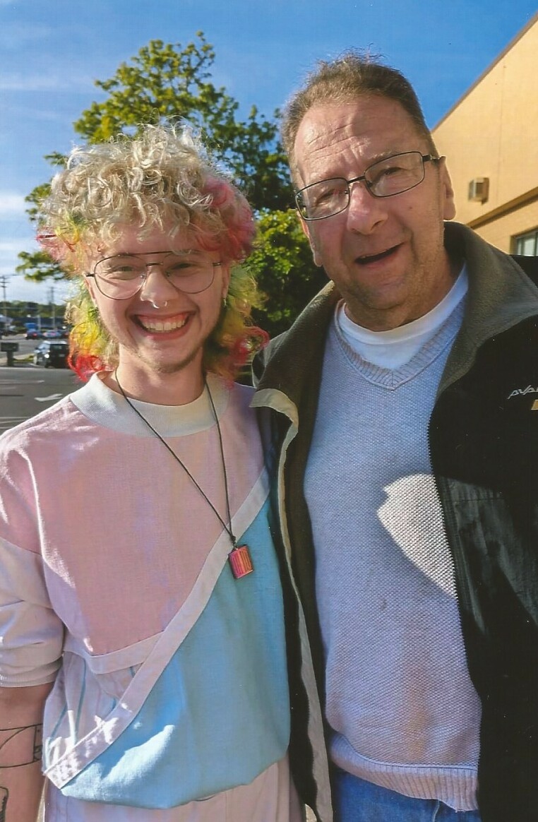 Two men posing together outdoors on a sunny day, both smiling at the camera near a beige building.