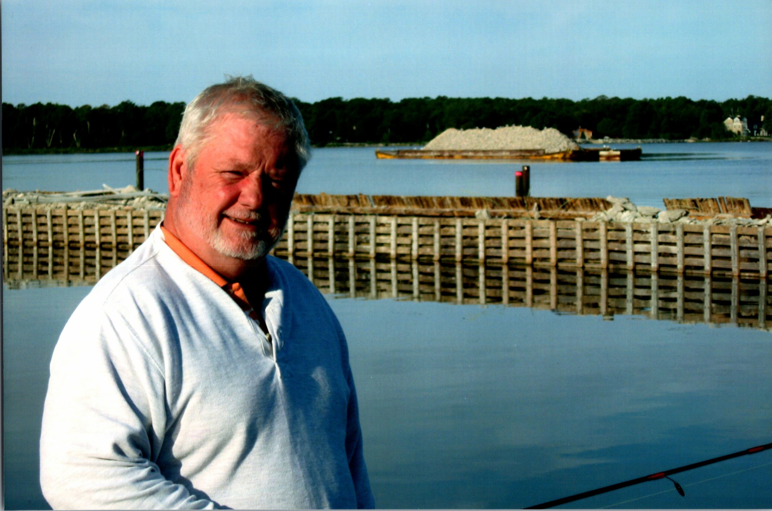 Man with gray hair and beard standing by a calm river, wooden retaining wall in the foreground and a gravel barge in the background.