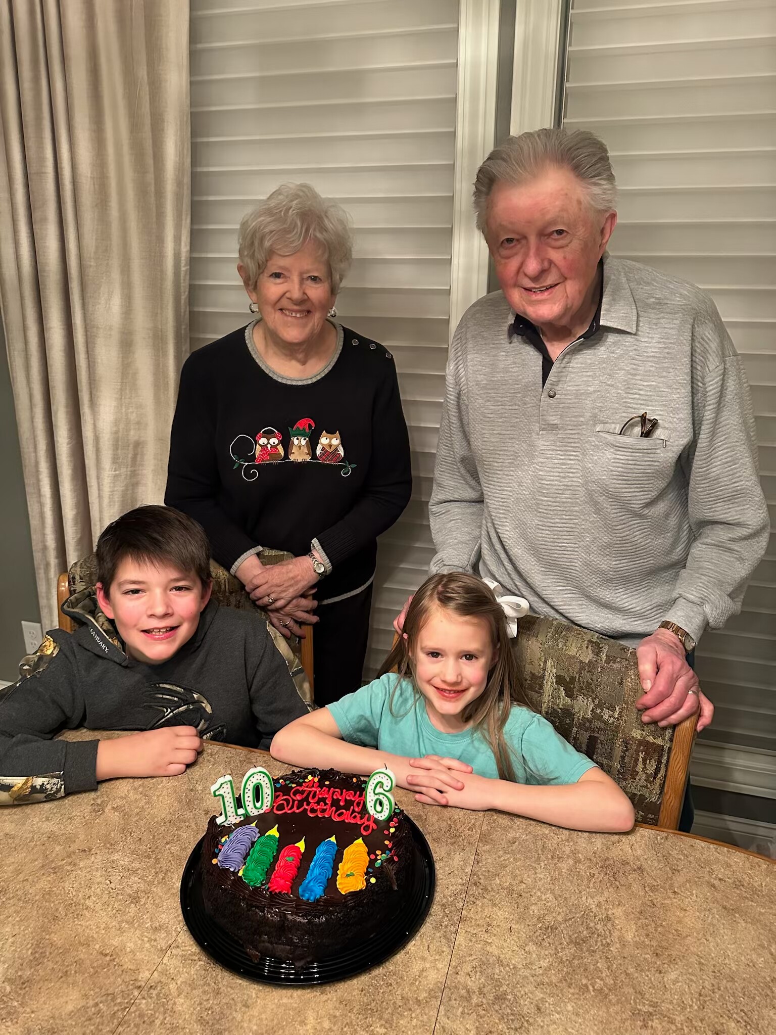 Two children sit at a table with a chocolate birthday cake in front of them, while two smiling grandparents stand behind them for a family birthday photo, cake decorated with colorful frosting and number candles '10' and '6'.