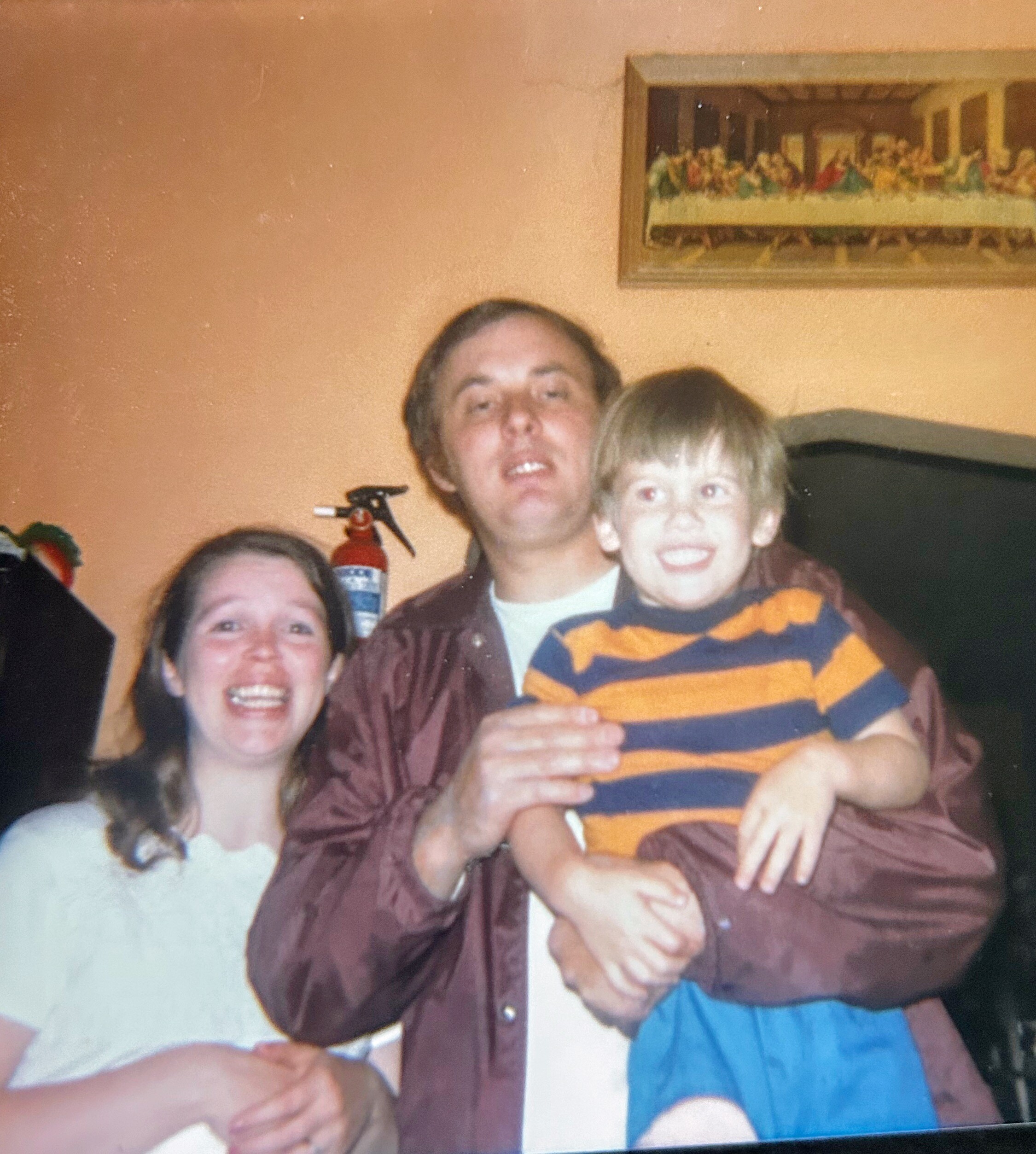 Smiling family group indoors: man holding a striped orange-and-blue shirted child with a woman beside them, in a room with an orange wall and a framed painting behind.