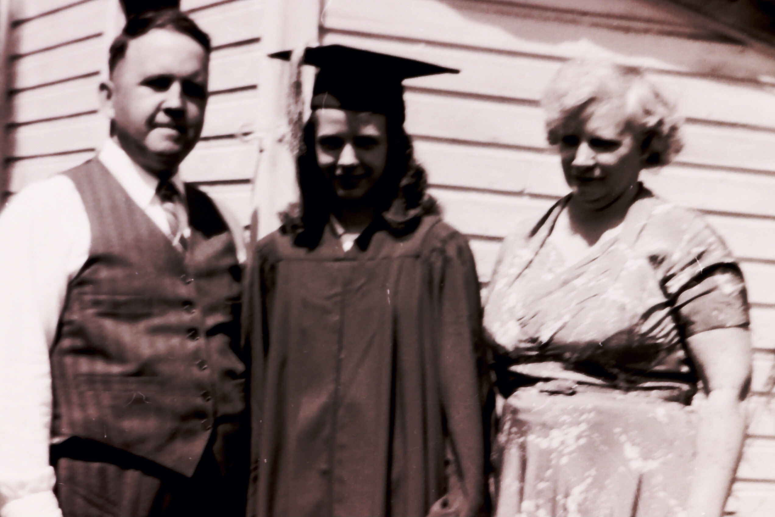 Three people posing for a graduation photo outdoors, the center person wearing a cap and gown while flanked by two others.