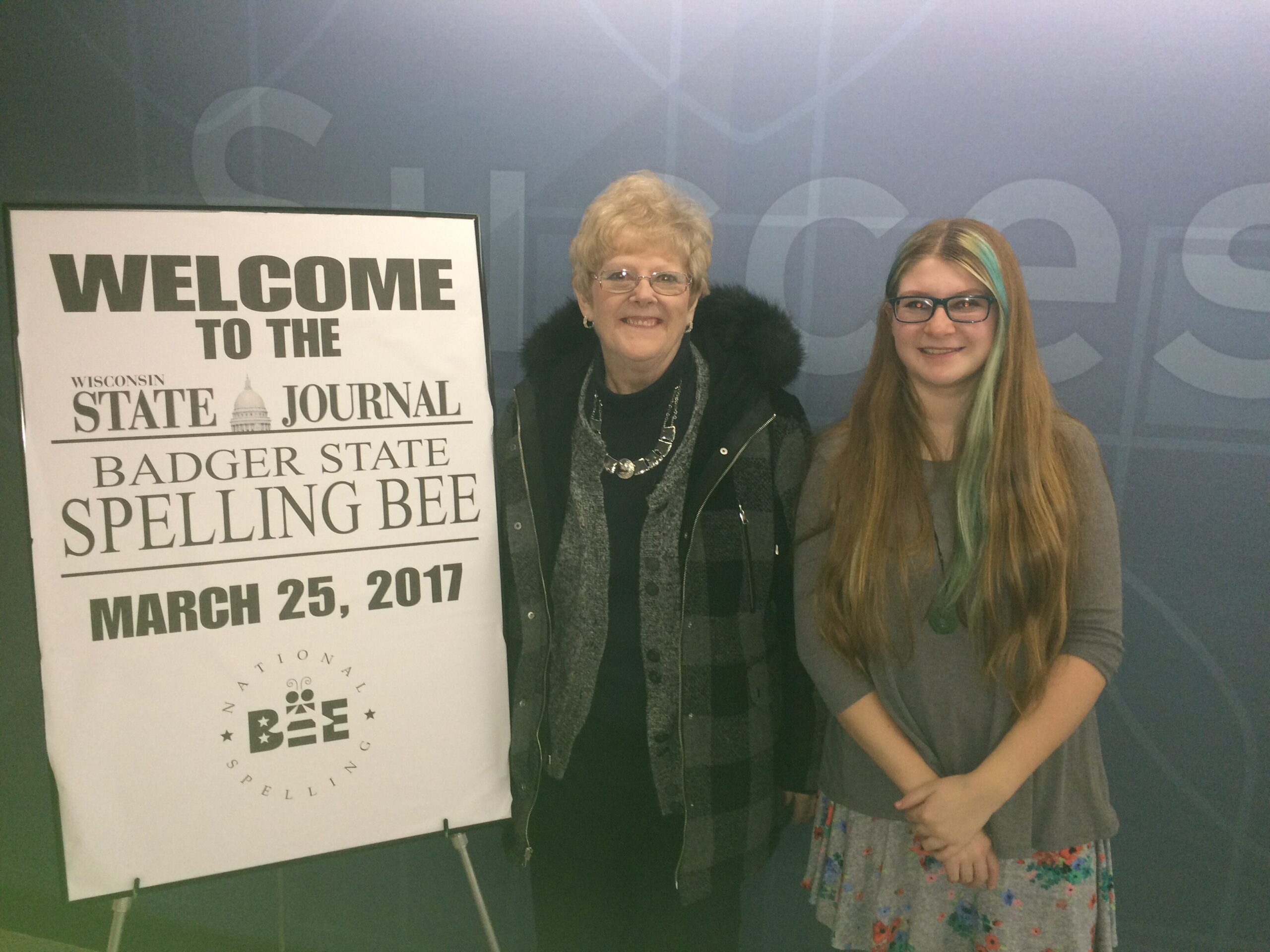 Two smiling women stand beside a large poster that reads 'Welcome to the Wisconsin State Journal Badger State Spelling Bee' at an event.