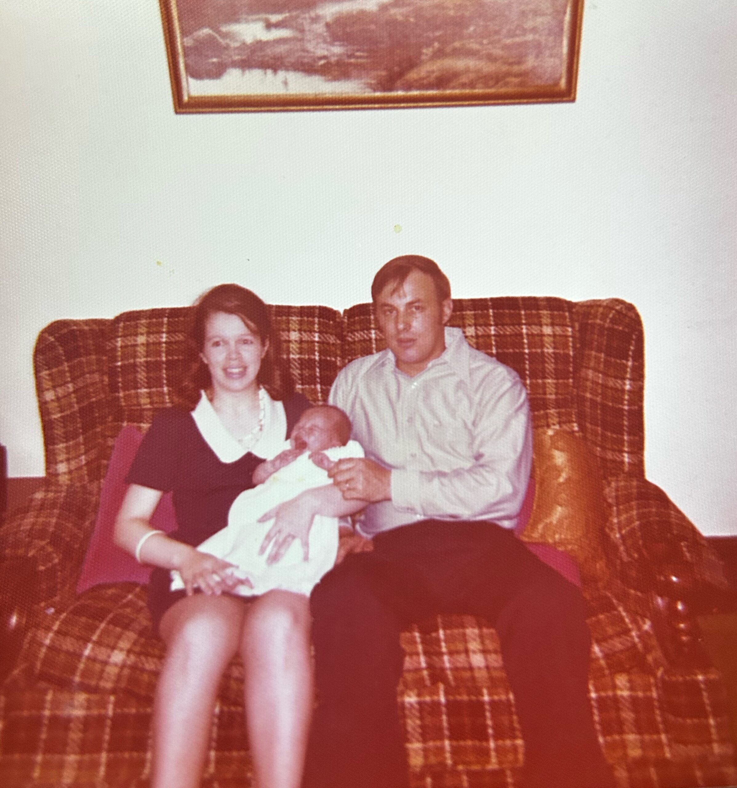 Couple seated on a plaid sofa, holding their newborn baby, with a framed painting above them.