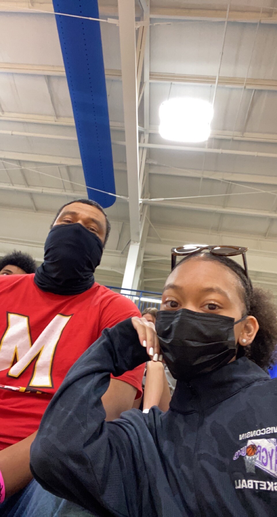 Two people wearing face masks take a low-angle selfie in a gym or indoor arena; a man in a red shirt with a large white-yellow M stands on the left, a woman in a black jacket with sunglasses on her head sits on the right.