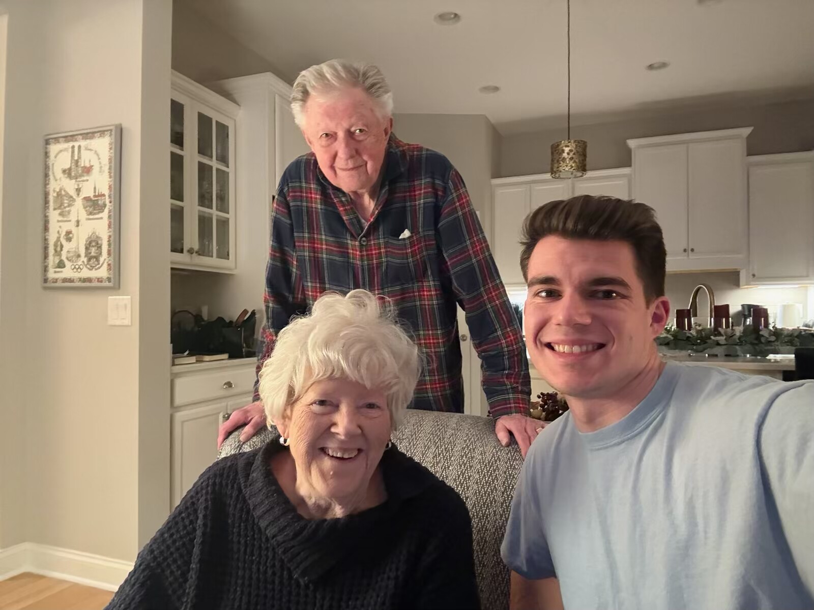 Three generations smiling for a selfie in a bright kitchen: a young man on the right, an elderly woman seated, and an older man standing behind them.