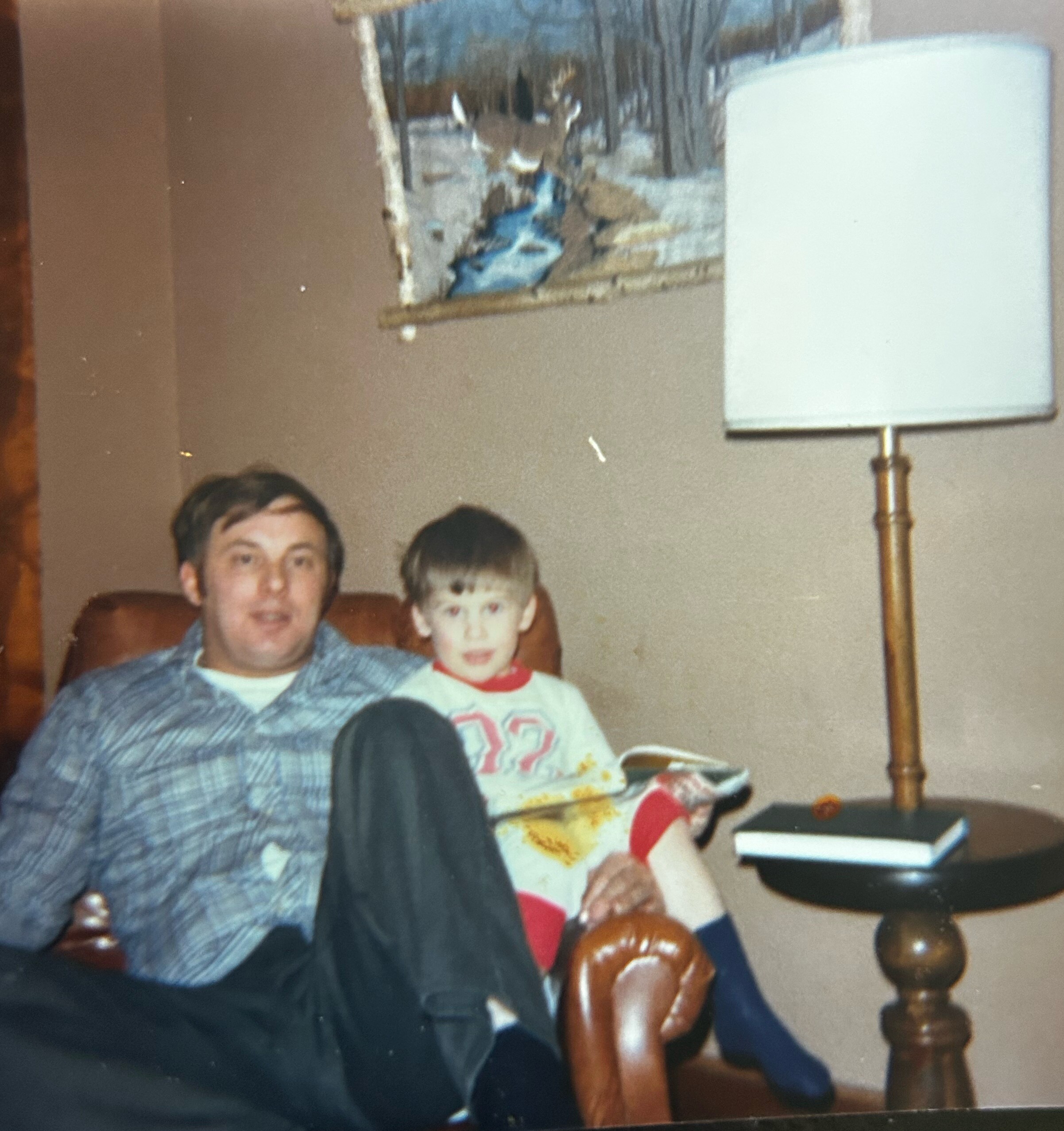 Man and boy seated together on a brown sofa in a living room, with a tall floor lamp to the right and a framed winter landscape on the wall behind them.