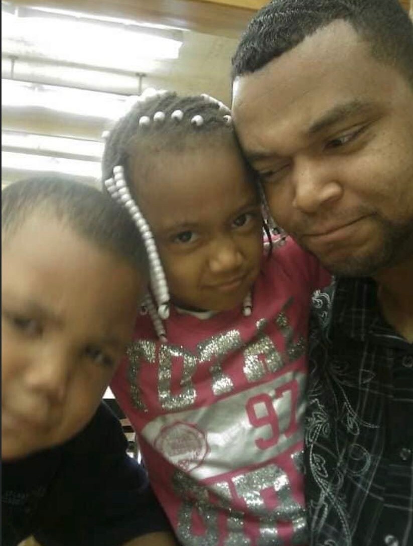 Man and two children pose for a selfie, all smiling at the camera; a girl with beaded braids rests her head on his shoulder while another child is nearby.