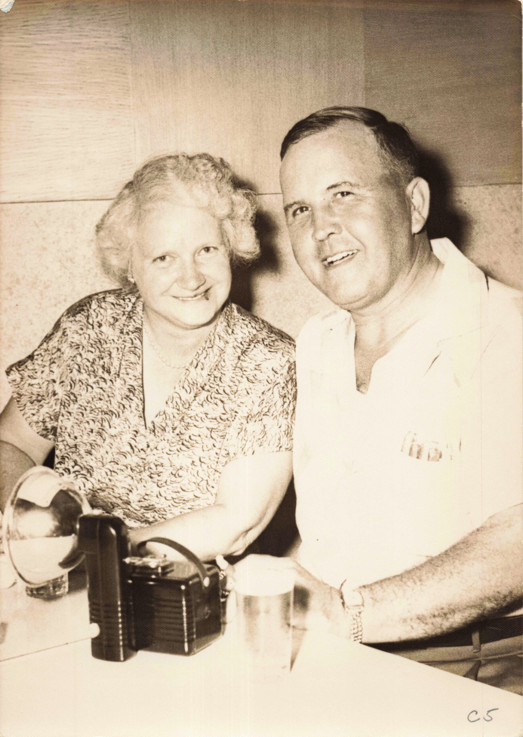 Smiling elderly woman and man seated at a table with a vintage camera in front of them on the table.