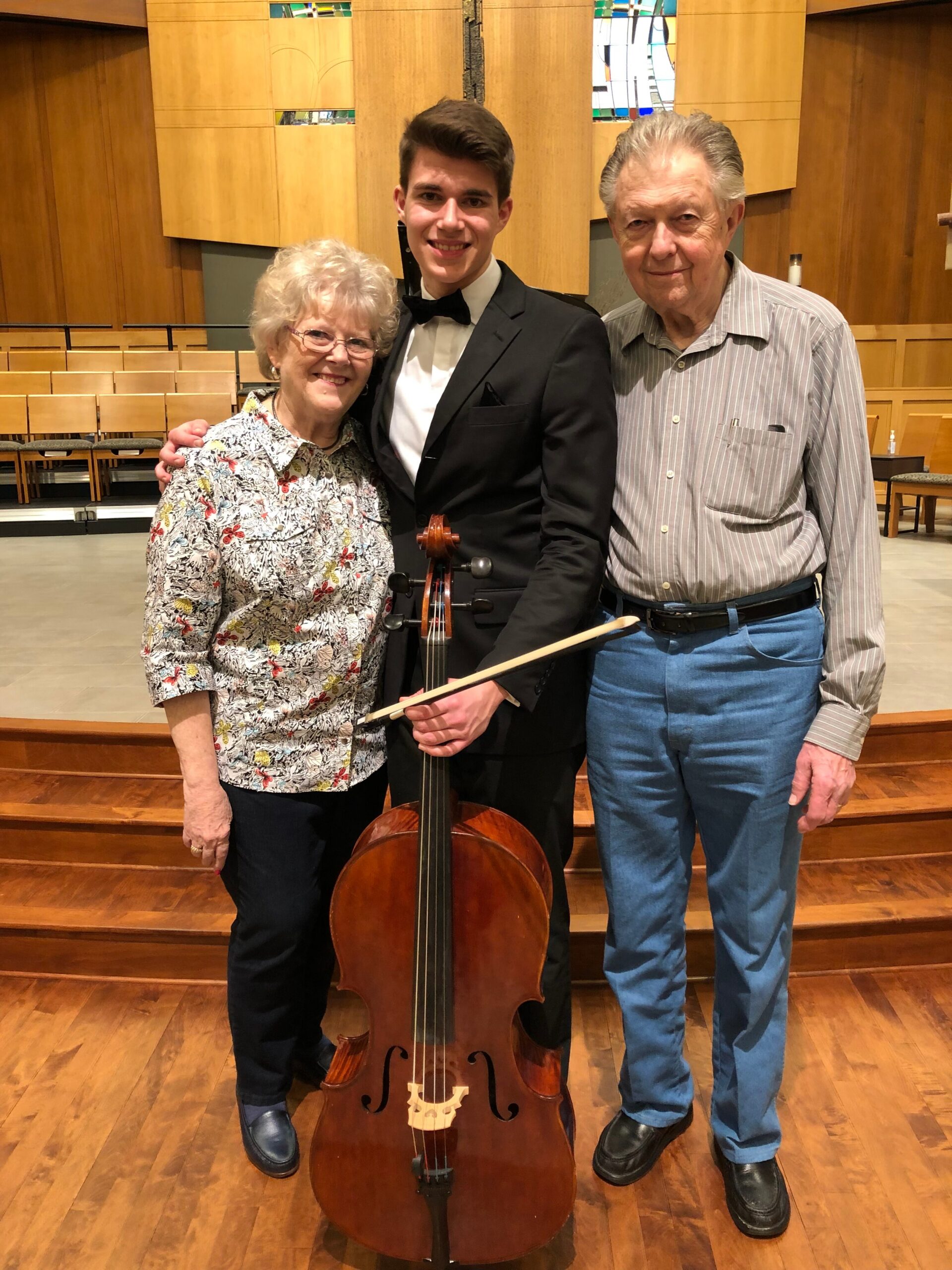 Three people pose with a cello on a wood-paneled concert hall stage: an elderly woman, a young cellist in a tuxedo, and an older man in a striped shirt smiling.