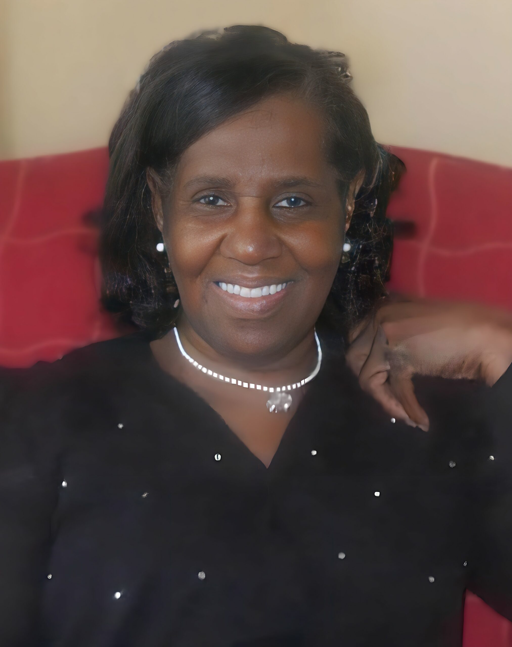 Close-up portrait of a smiling woman with dark hair, wearing a pearl necklace and earrings, seated on a red couch against a beige background.