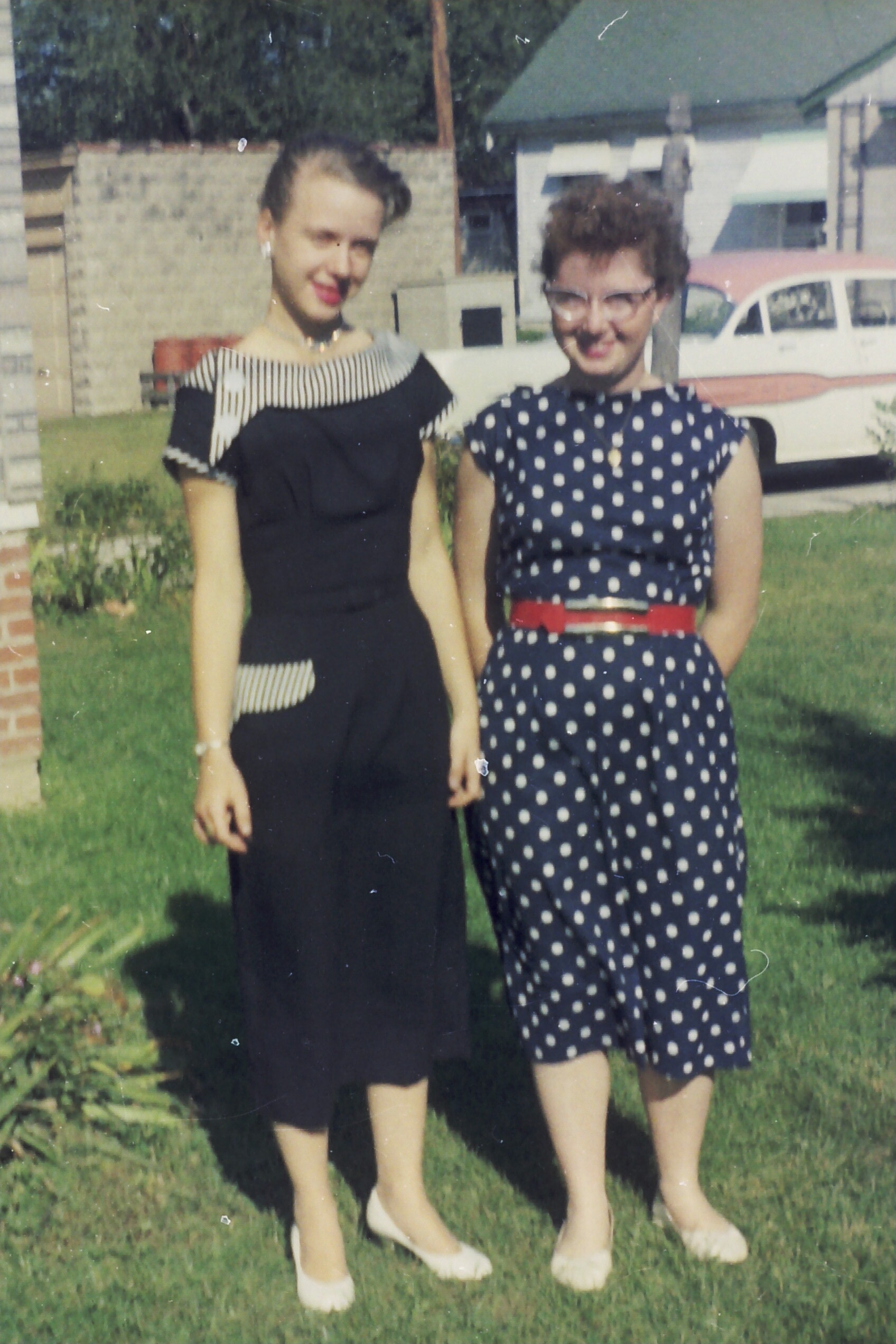 Two women stand on a lawn in vintage dresses: one in a black dress with striped shoulders, the other in a navy polka-dot dress with a red belt.
