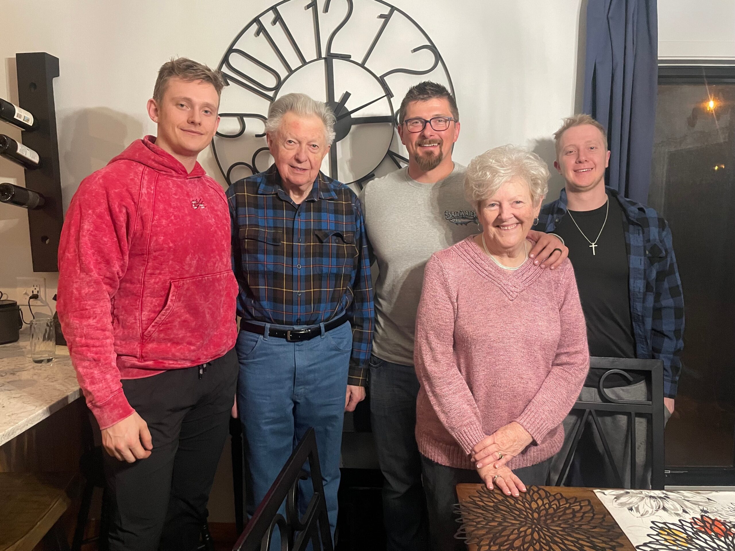 Five people standing together and smiling for the camera in a home dining area with a large clock on the wall behind them.