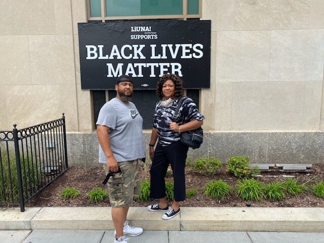 Two people stand on a sidewalk in front of a Black Lives Matter sign on a building; man in a gray T‑shirt and camo shorts, woman with a backpack.