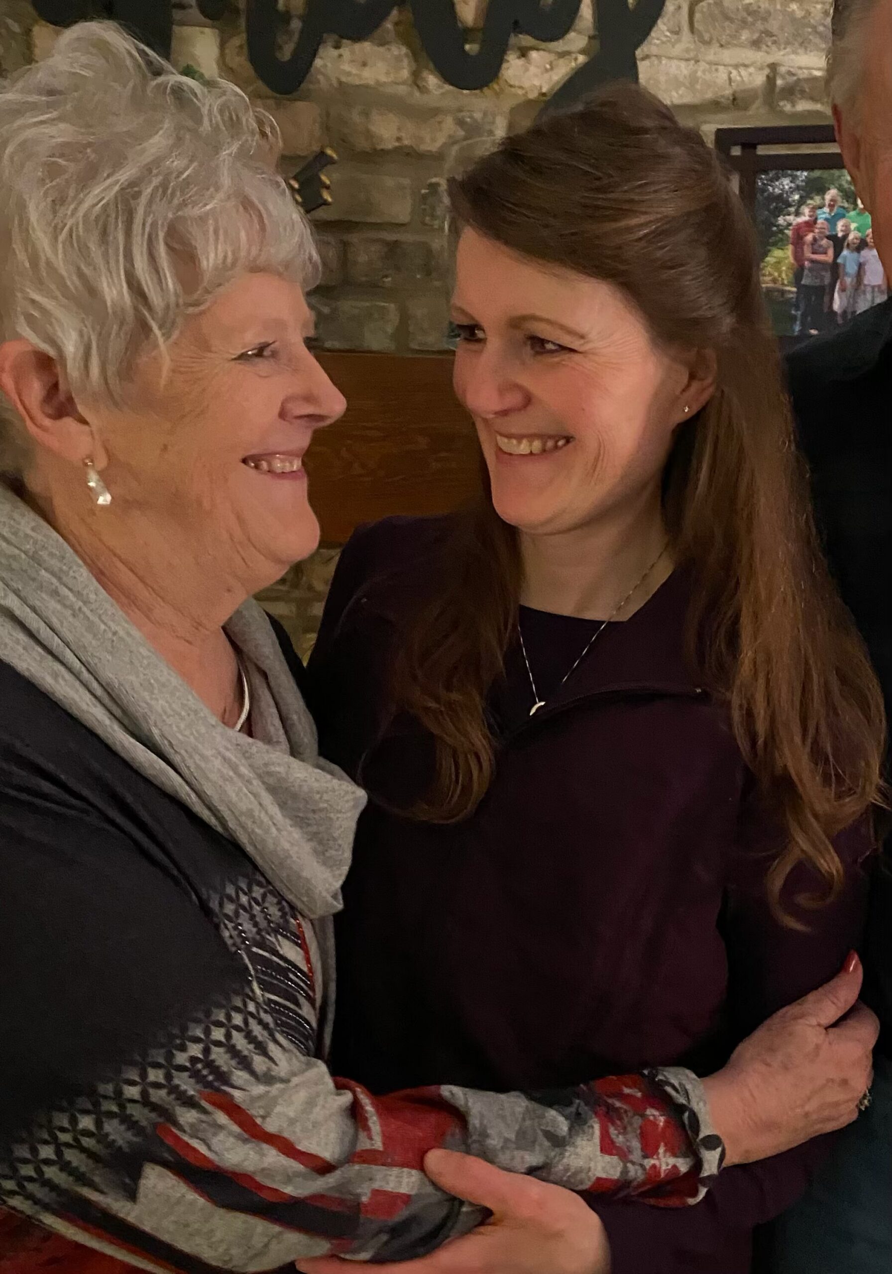 Two women share a warm hug and smile at each other in a cozy indoor setting with a stone wall behind them, showing affection.