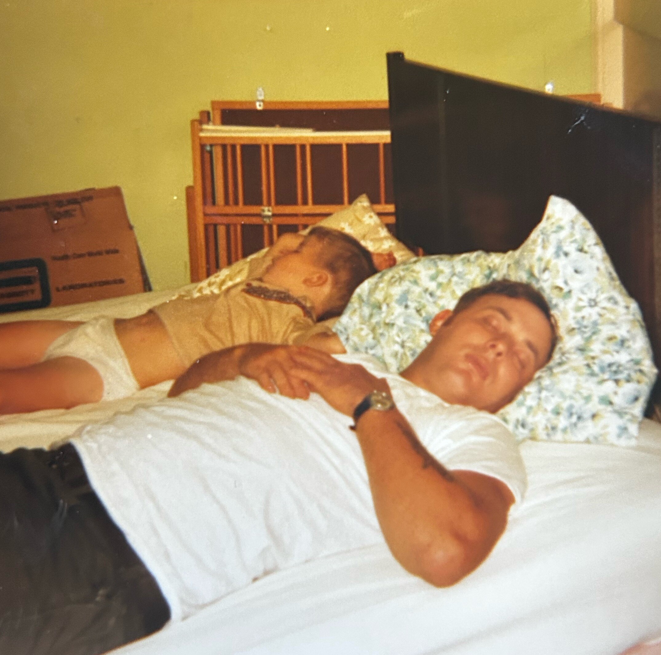 People lying on a bed under blankets in a dim room, drifting asleep beside a dark wall and wooden headboard backdrop