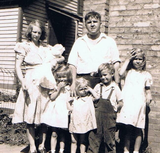 Old black-and-white photo of six children and two adults standing outside a house, posing for a family portrait.