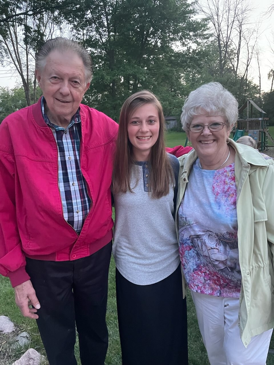 Three people pose together outdoors: an elderly man in a pink jacket, a smiling teenage girl, and an elderly woman in a light jacket in a grassy park area.