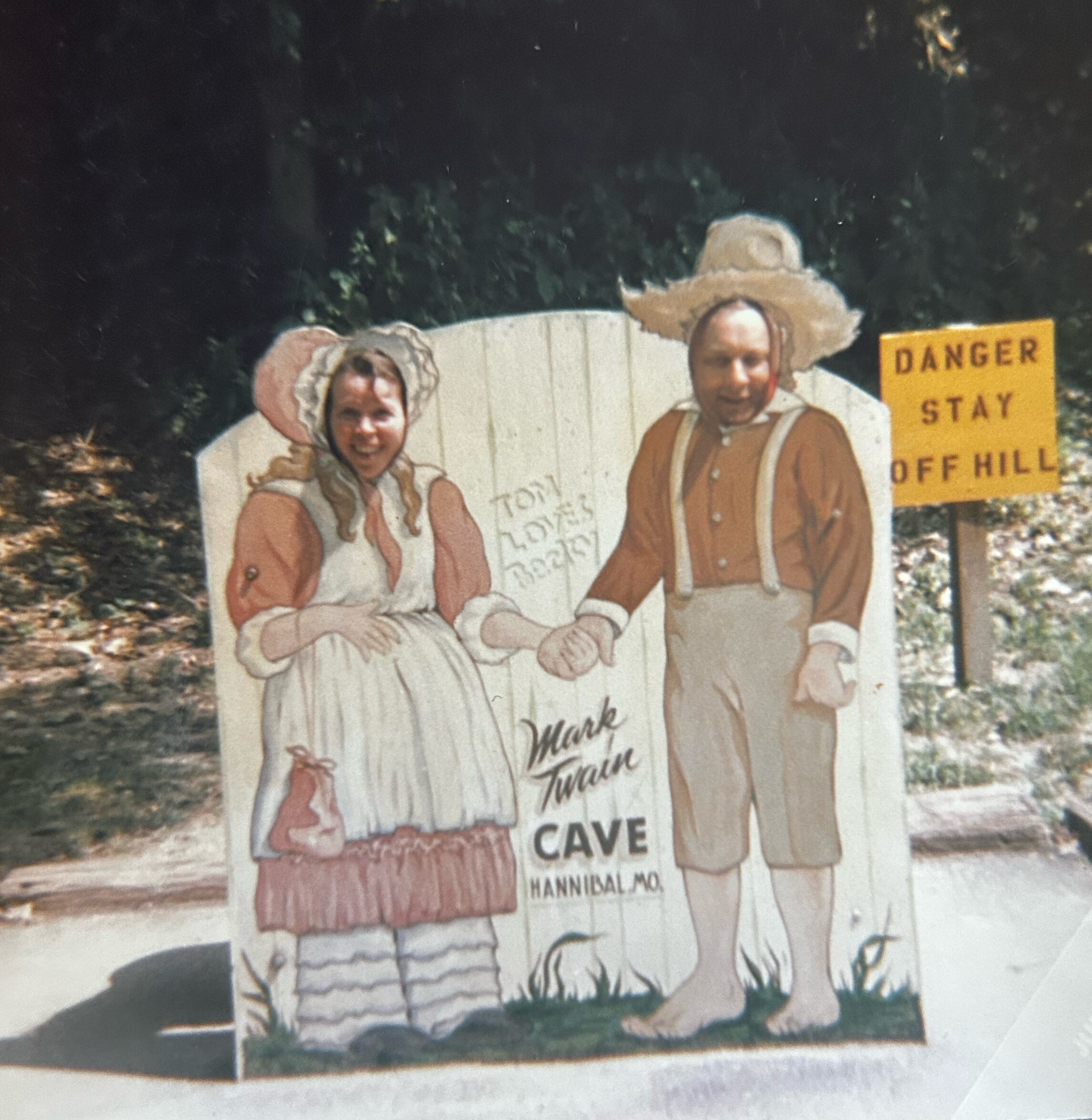 Two people in cartoonish animal costumes posing beside a yellow danger sign outdoors on a white display board