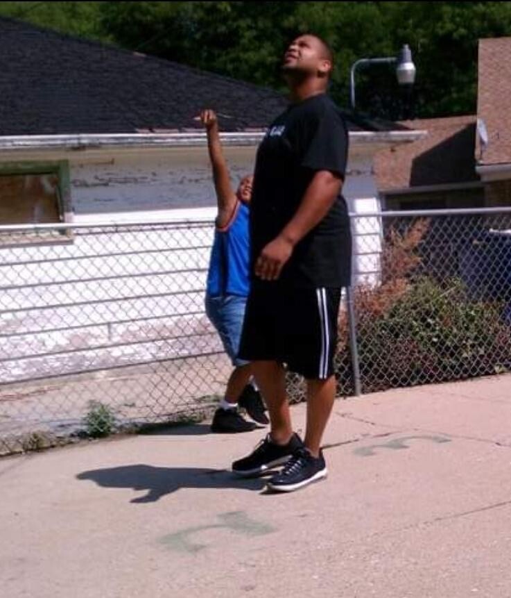 Two men outdoors near a chain-link fence; one in a black t-shirt looks upward while a boy in blue behind him raises an arm.