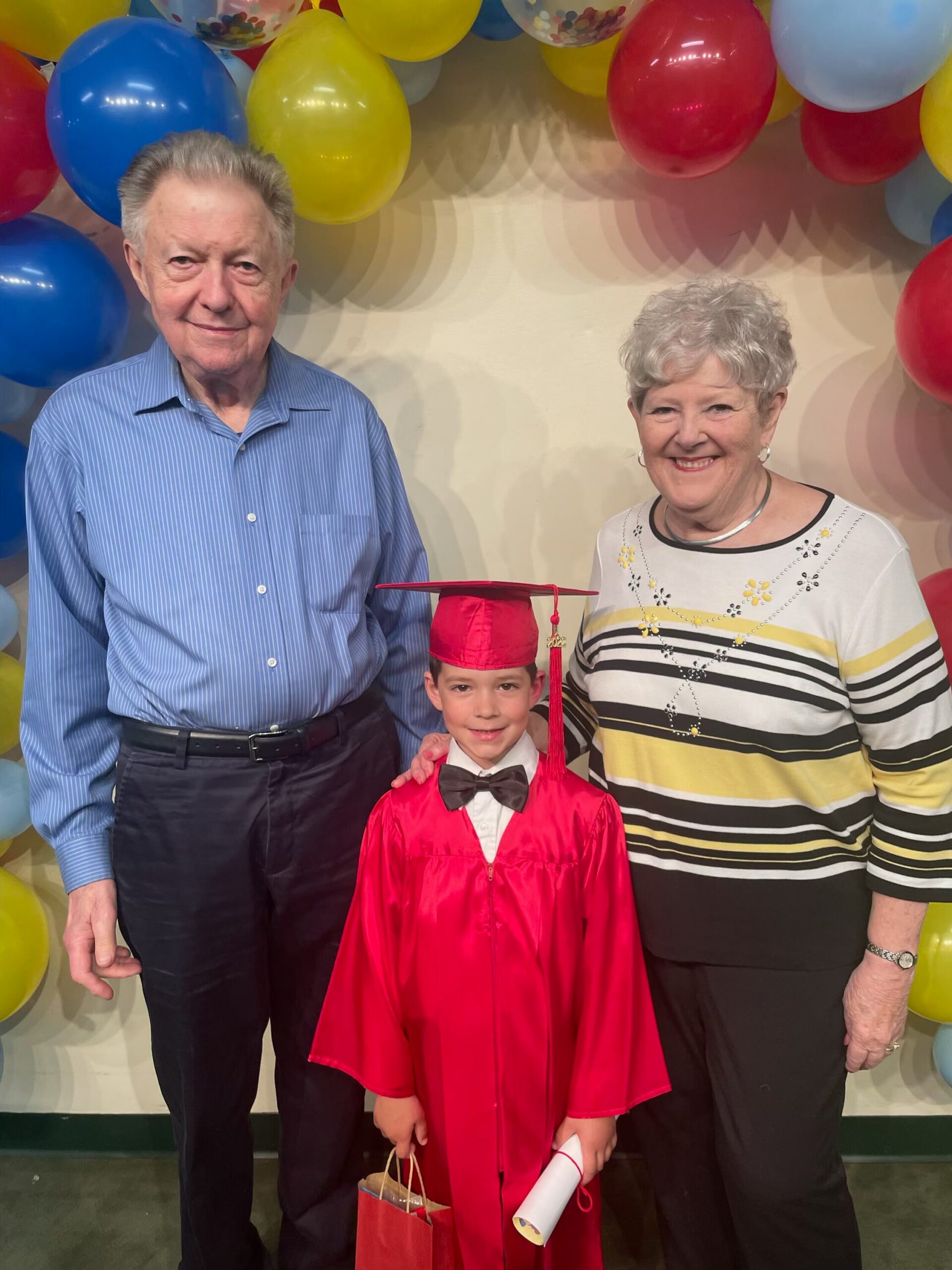 Young graduate in red cap and gown with diploma, flanked by his grandparents under colorful balloons.