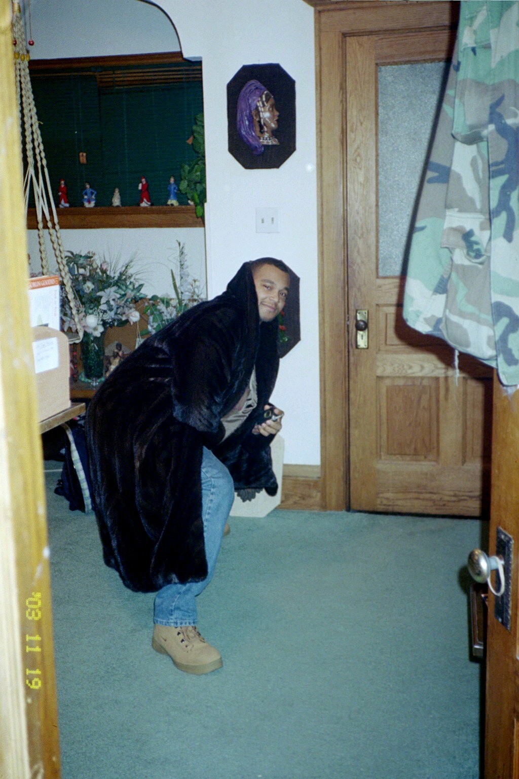 Person in a black hooded cloak crouches in a doorway, looking toward the camera in a cluttered room.