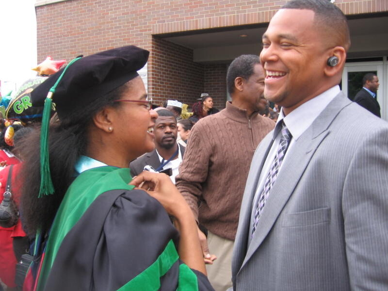 Graduate in cap and gown smiling with a man in a gray suit outside a brick building at a graduation celebration.