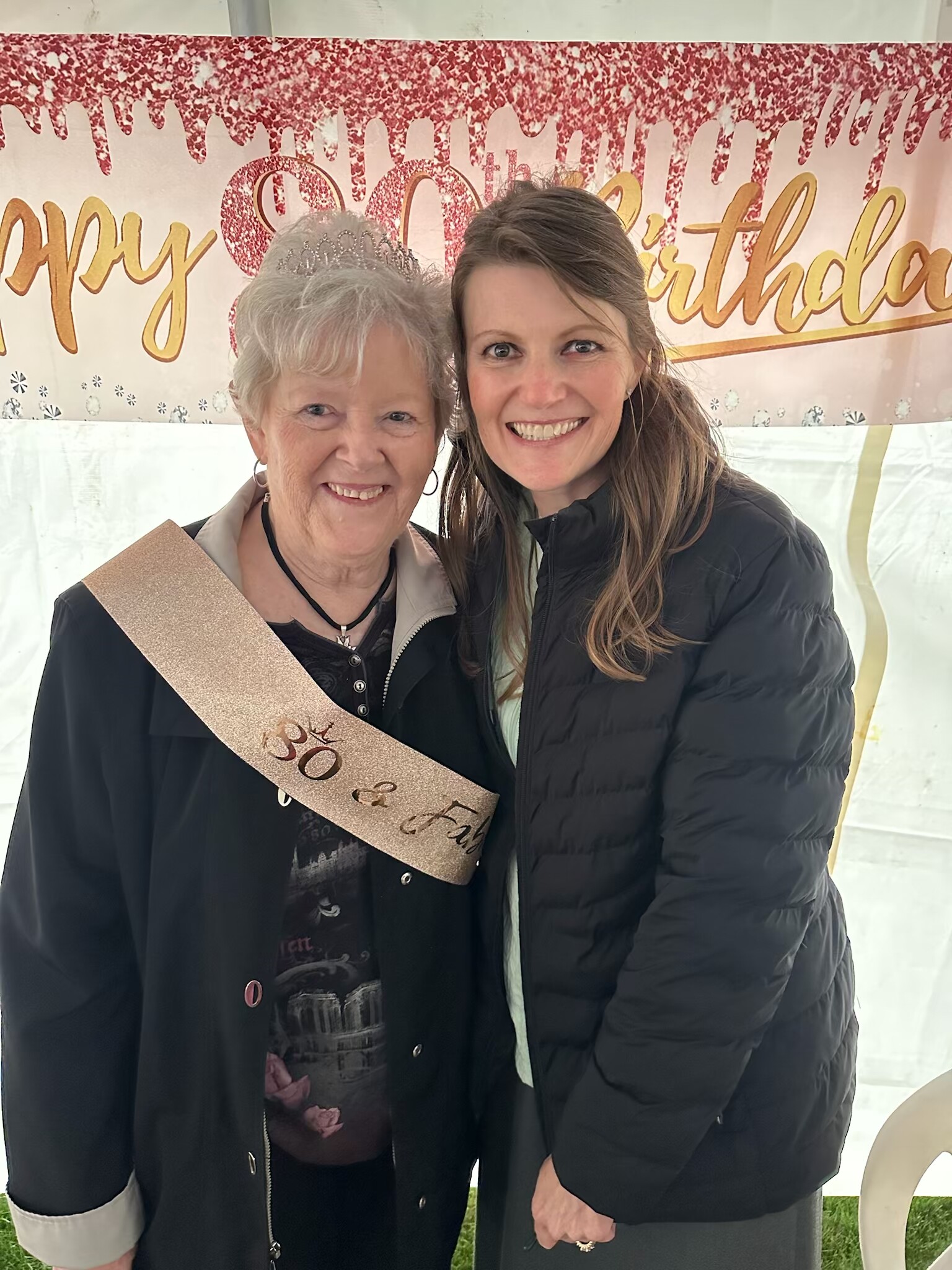 Two smiling women pose at a birthday party in front of a glittery pink “Happy Birthday” backdrop; the older woman wears a tiara and sash.