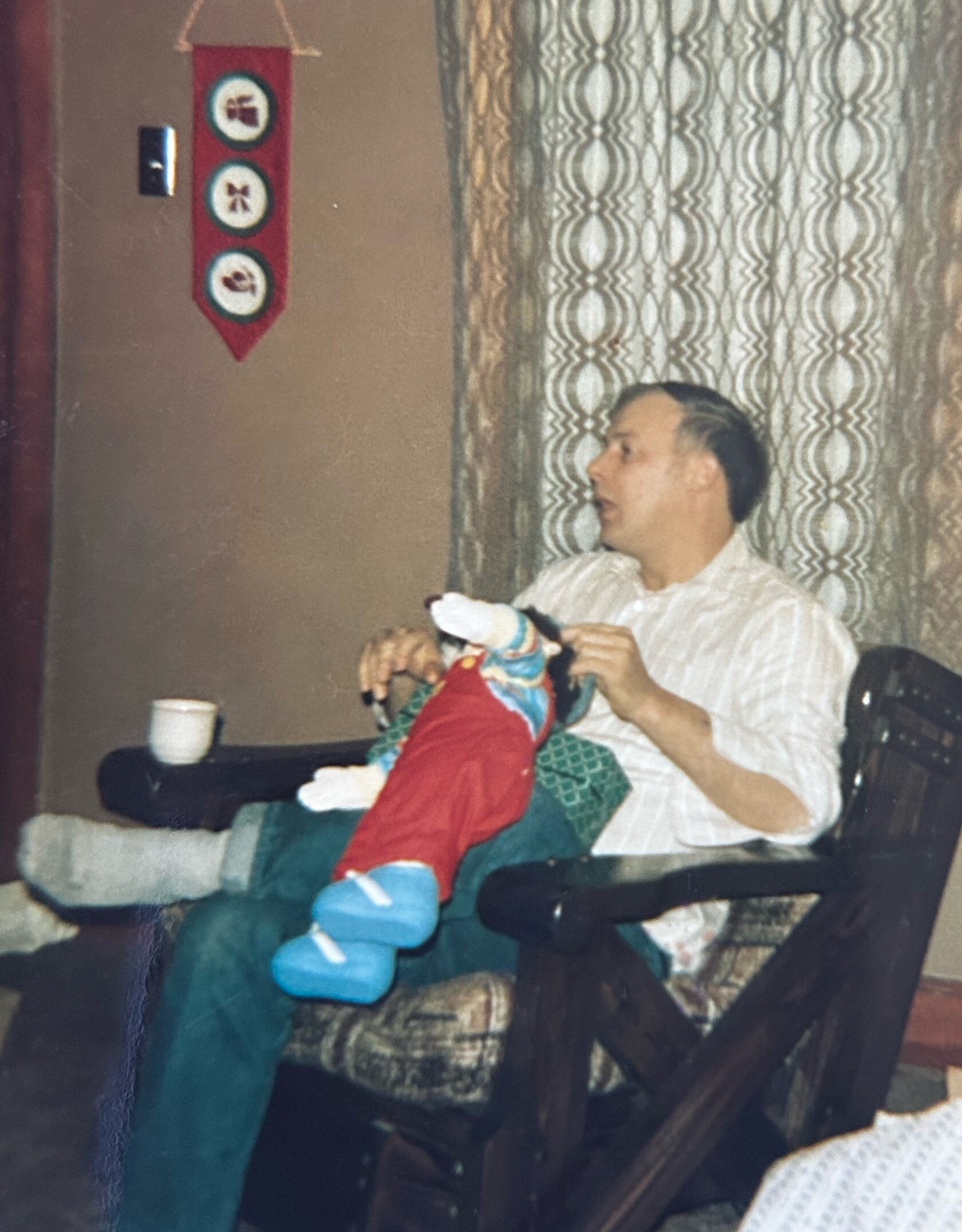Man seated in a living room, holding a colorful puppet, wearing a striped shirt; patterned curtains behind him.