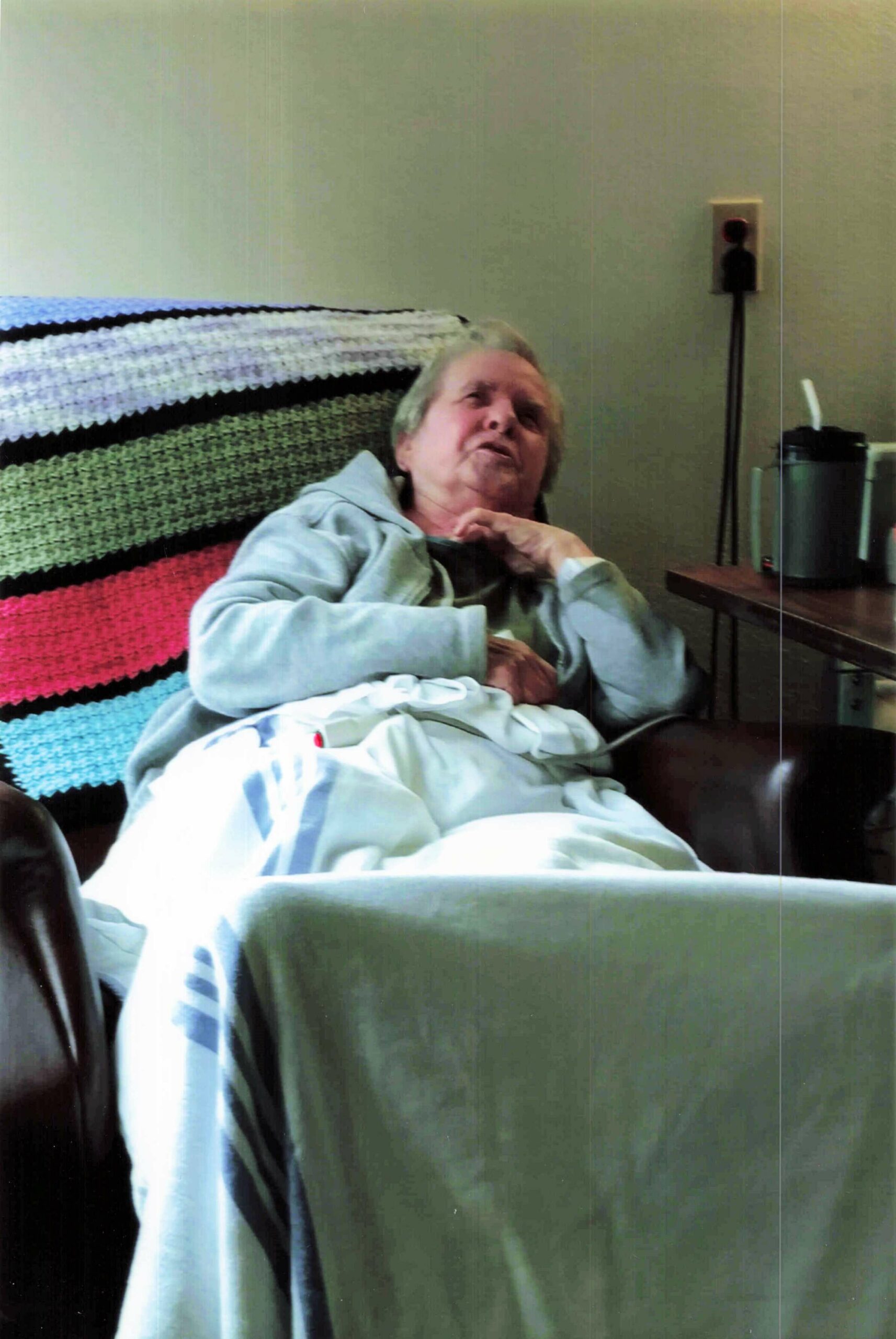 Elderly person resting in a hospital bed, covered with a light blue blanket, hands folded on chest.