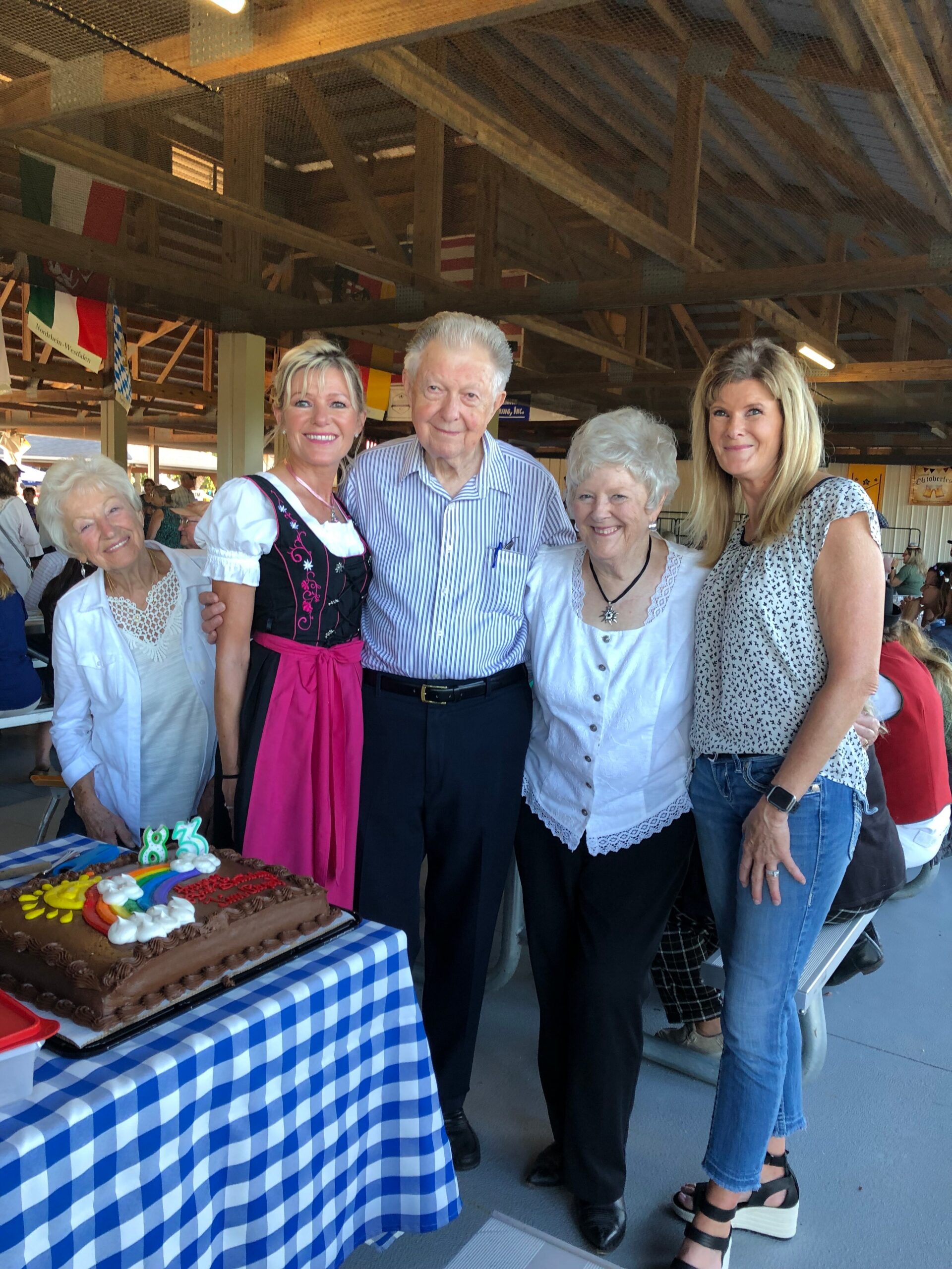 Five adults pose together beside a large chocolate cake with rainbow decorations on a blue-and-white checkered tablecloth at a party under a wooden pavilion as flags hang overhead:**