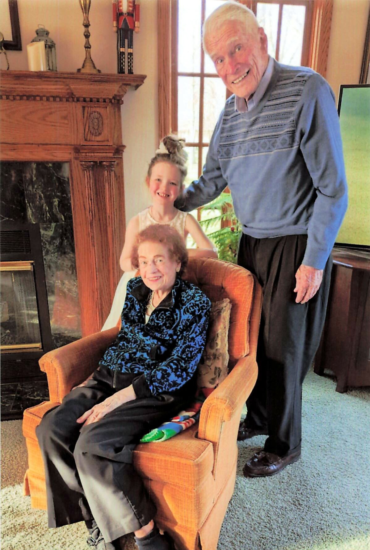 A smiling elderly couple with a granddaughter standing behind them in a cozy living room by a fireplace.