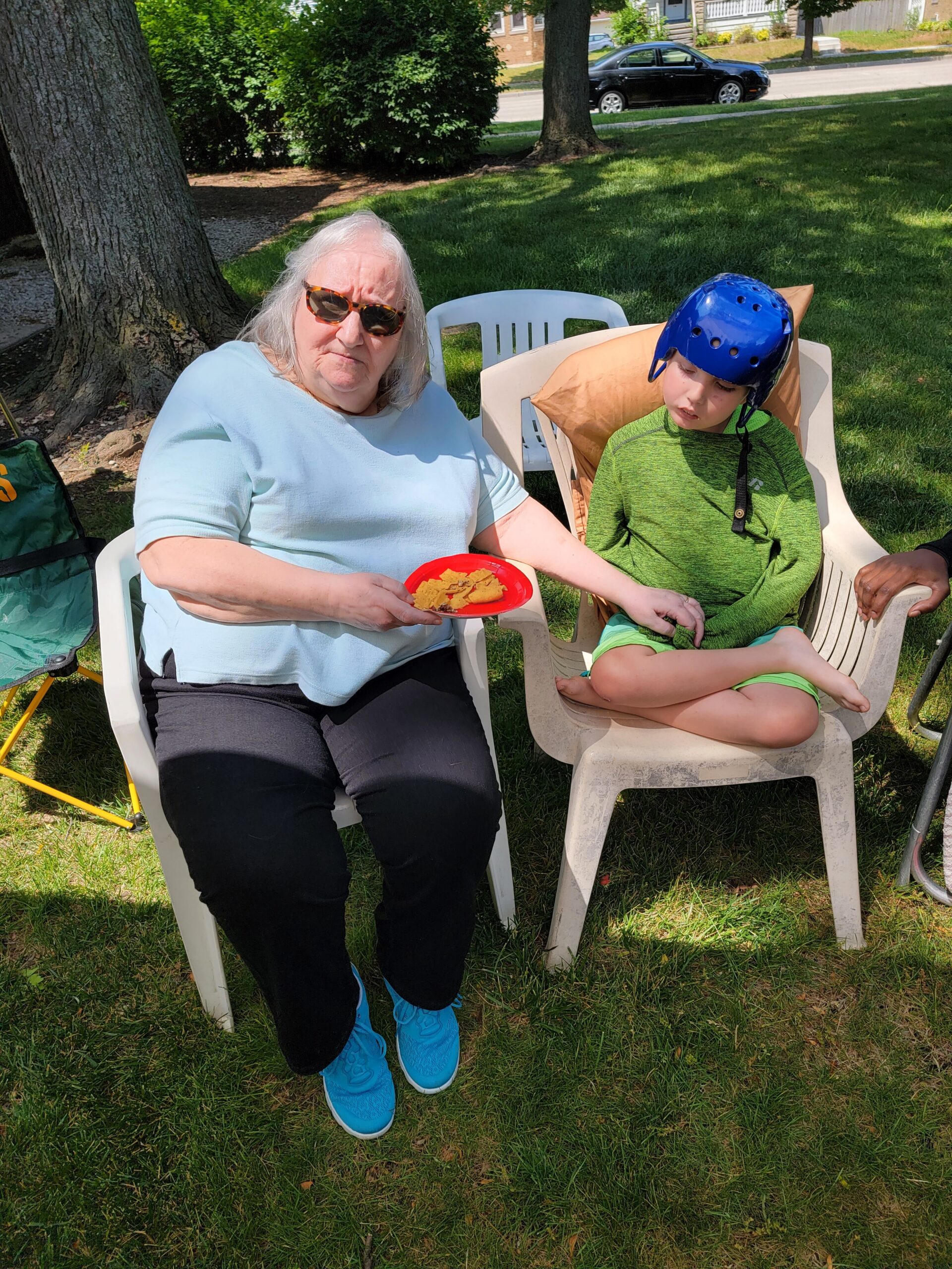 Senior woman with sunglasses sits in a white plastic chair outdoors, holding a red plate of crackers while a young boy wearing a blue helmet sits nearby on the grassy yard.