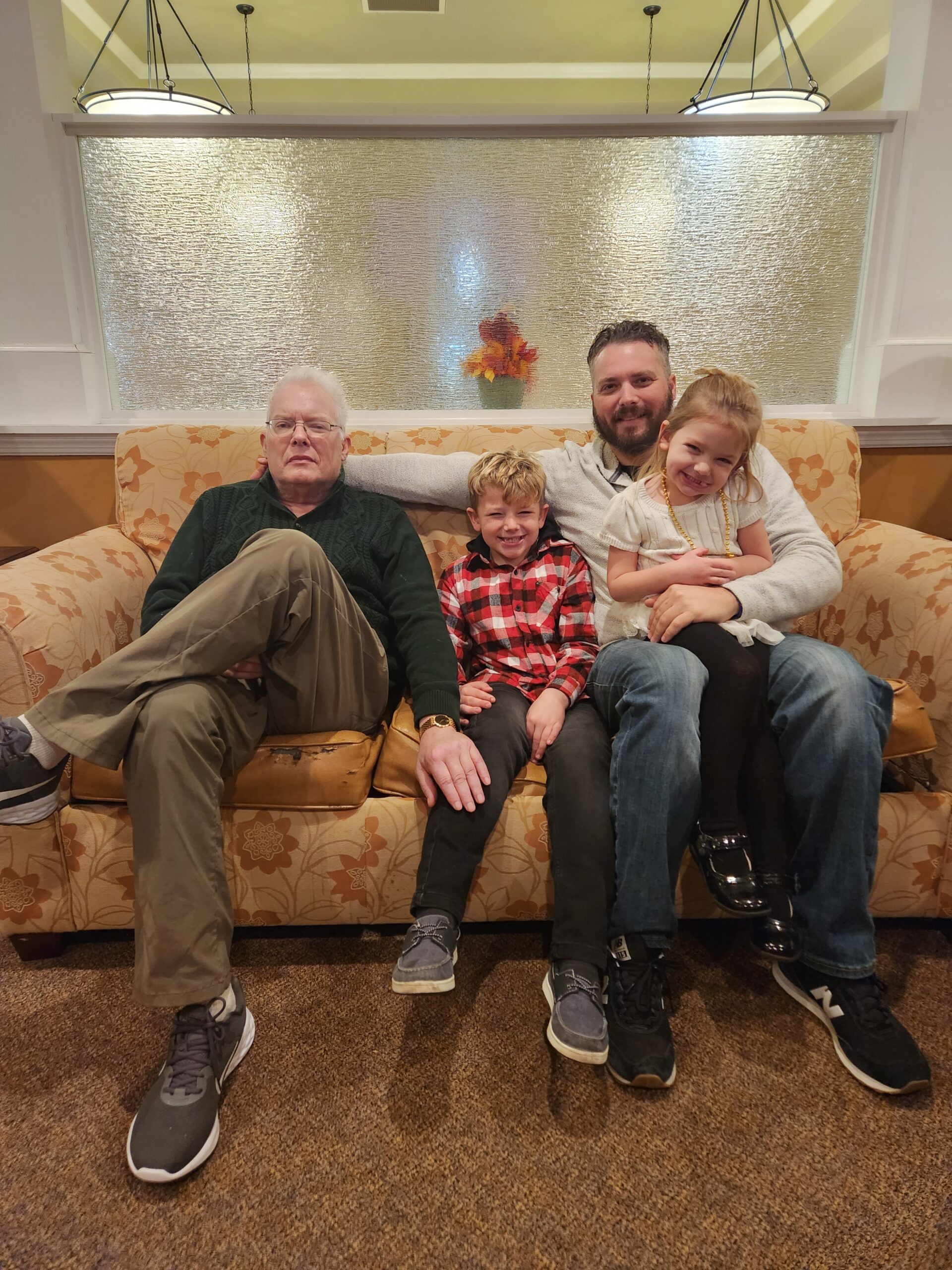 Four people sit on a beige floral couch in a living room: an older man on the left, a smiling boy in plaid, a bearded man, and a young girl in white.
