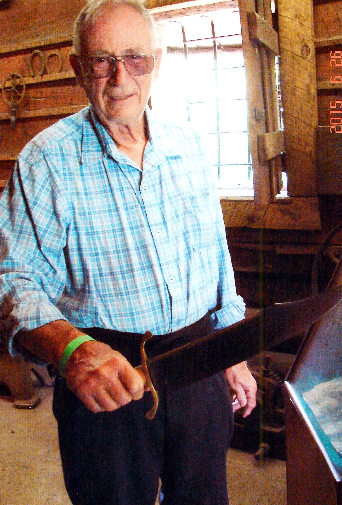 Older man with white hair and glasses in a blue plaid shirt stands in a rustic wooden workshop, holding a curved brass-handled tool.