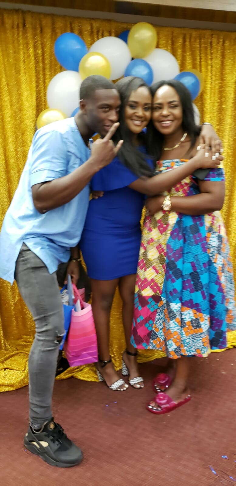Three friends pose together at a celebration, smiling with arms around each other in front of gold balloon backdrop and blue/gold balloons.