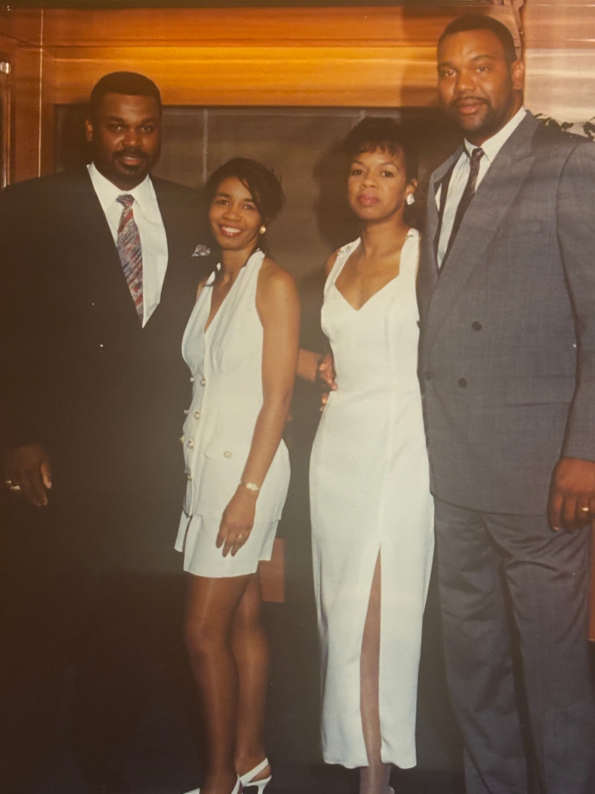 Group of four adults in formal attire posing for a photo indoors; two men in suits and two women in white dresses stand close together, smiling.