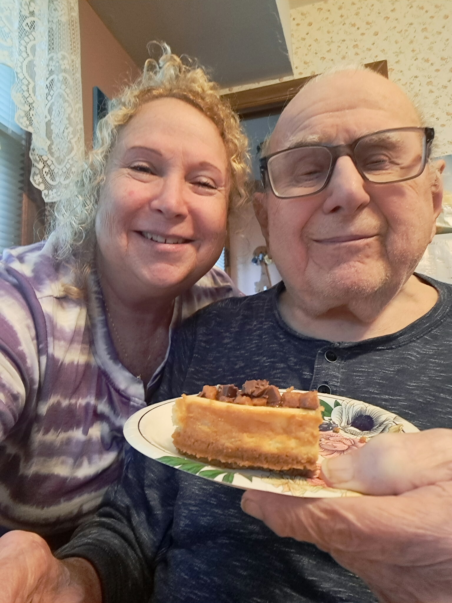 Smiling couple close up, woman on left and man with glasses on right, holding a plate with toast topped with meat in a kitchen setting.